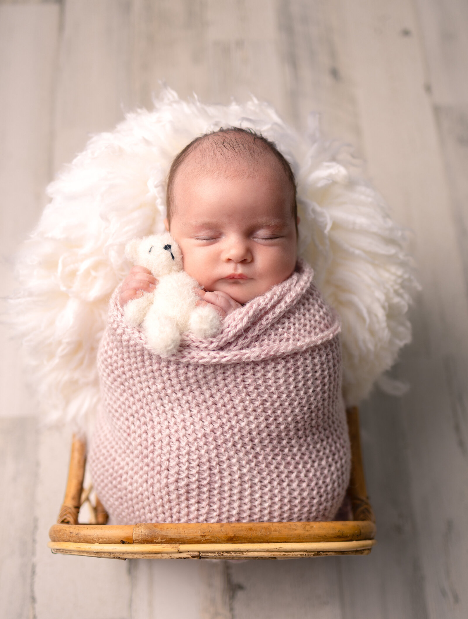 Newborn baby girl swaddled in a pink knit wrap holding a tiny white teddy bear during a professional studio session in Bergen County
