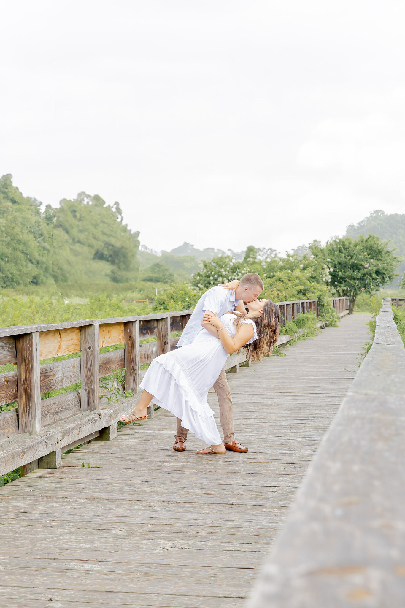 A man dips his wife on a wooden bride in Alexandria, Virginia. She's wearing a flowy white dress and he matches.