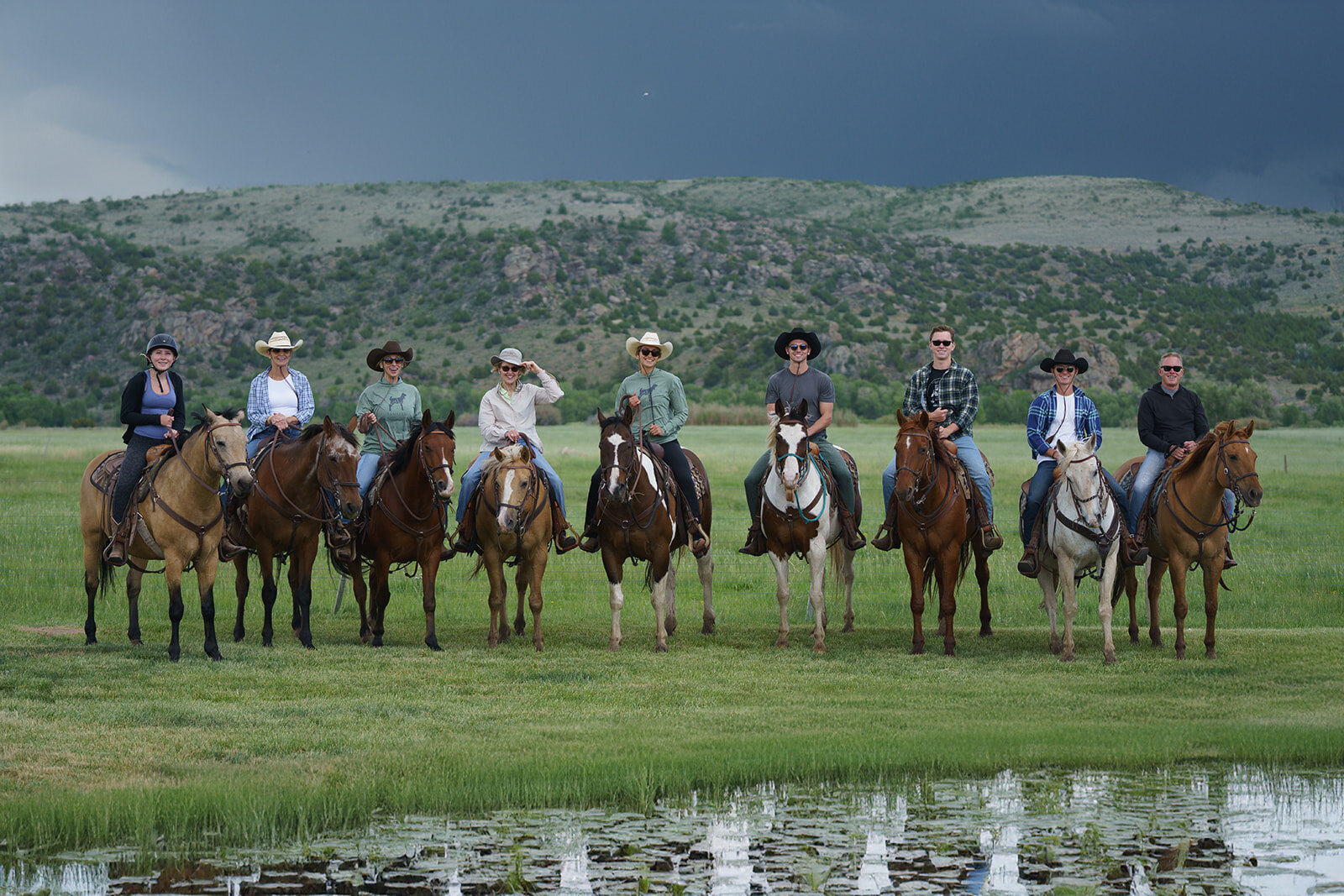Nine cowboys and cowgirls on horses with mountains and dark clouds in the background