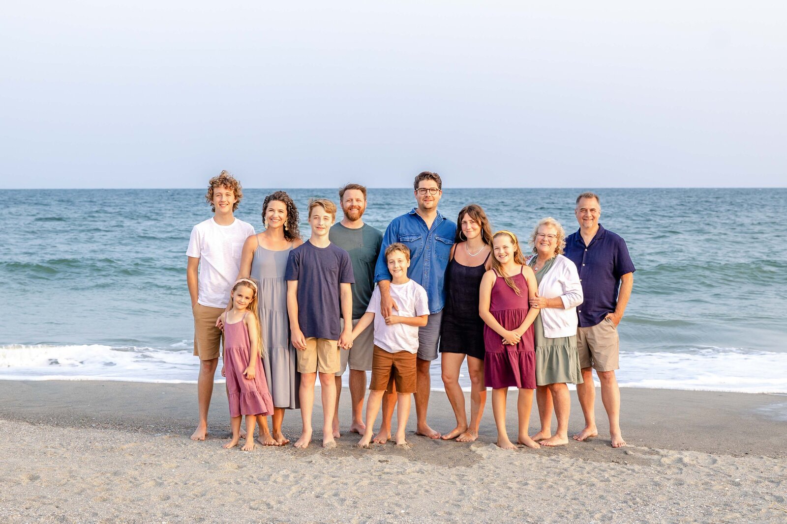 An extended family poses together on the beach, dressed in casual summer attire. The ocean waves and soft evening light create a serene backdrop, ideal for North Carolina family photography, capturing relaxed and joyful family moments.