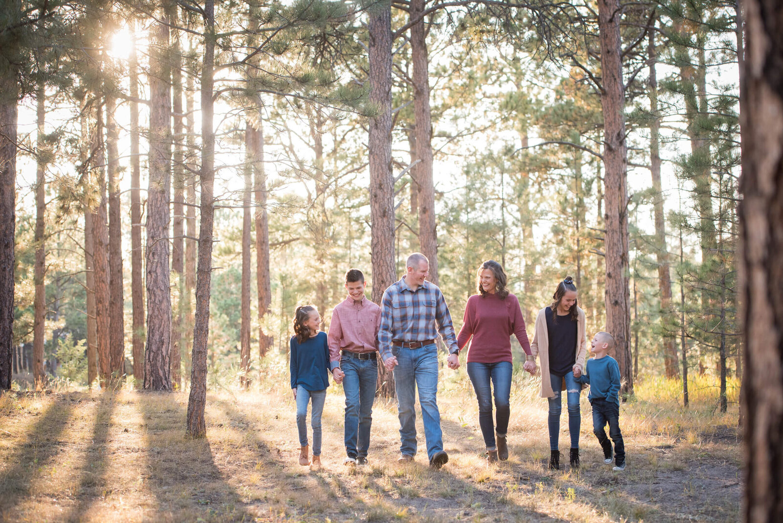 A family of 6 walks through a pine forest at sunset holding hands and laughing with each other