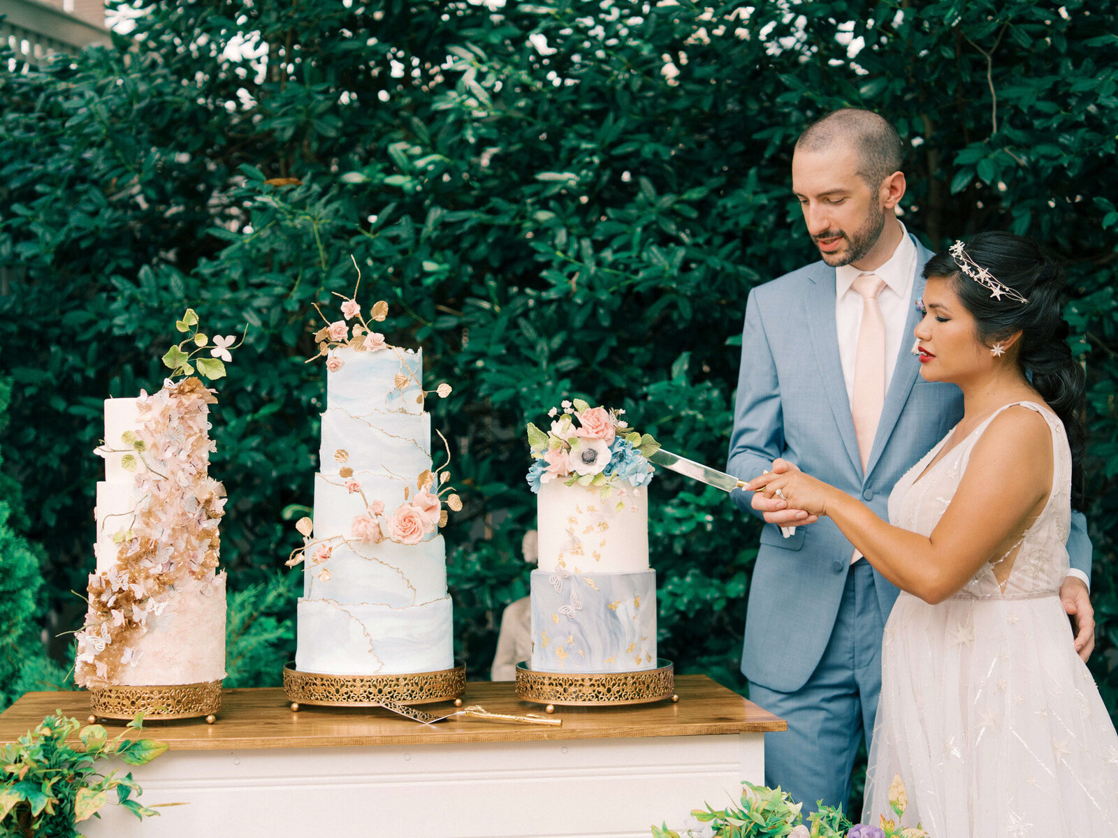 bride and groom standing to the right of a cake table with three luxury colorful two and three tiered cakes to cut the wedding cake 