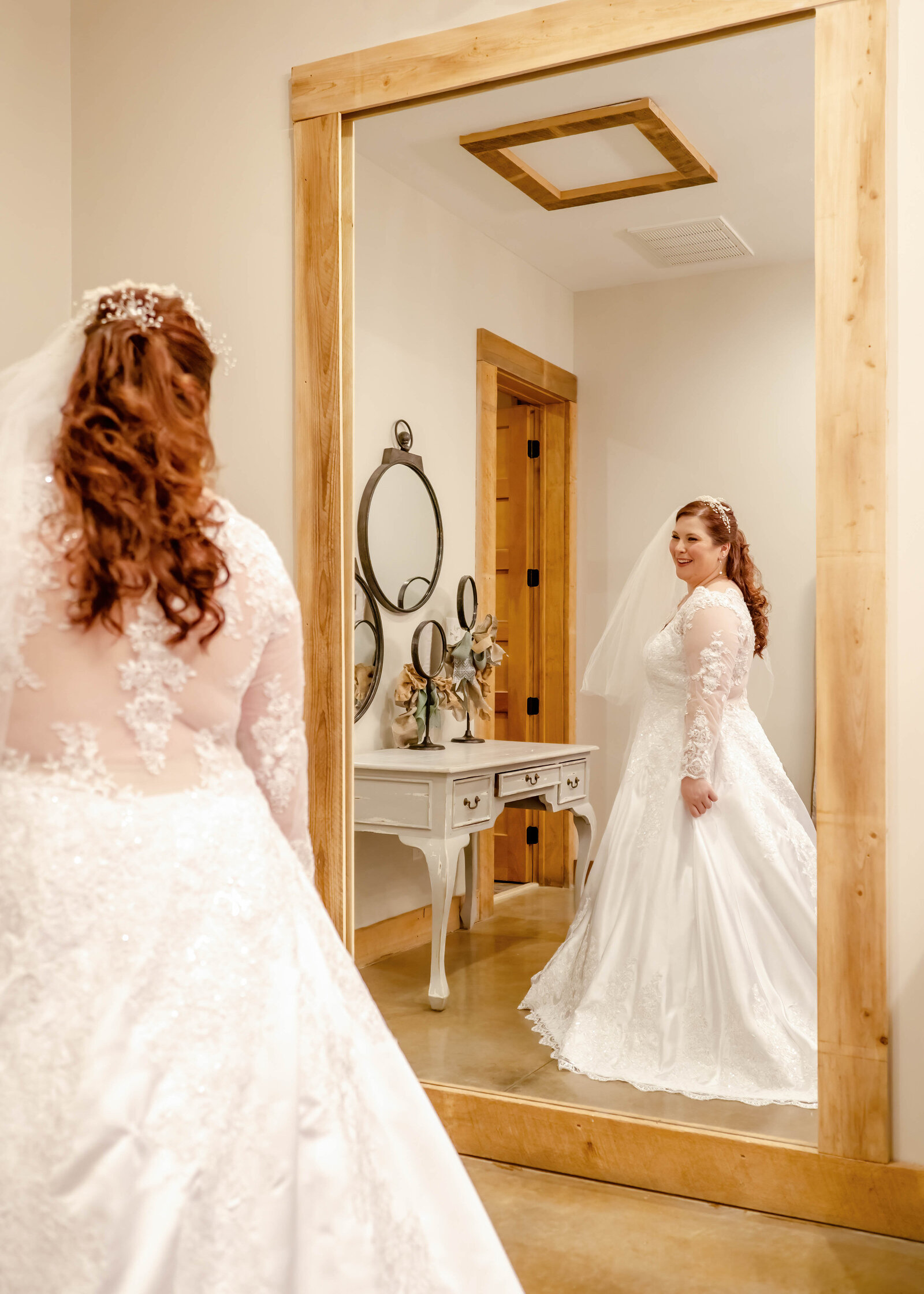 A bride in a lace wedding gown admires her reflection in a full-length mirror in a rustic bridal suite during a Dupline Winery, Wallace, NC wedding, capturing timeless elegance and authentic moments.