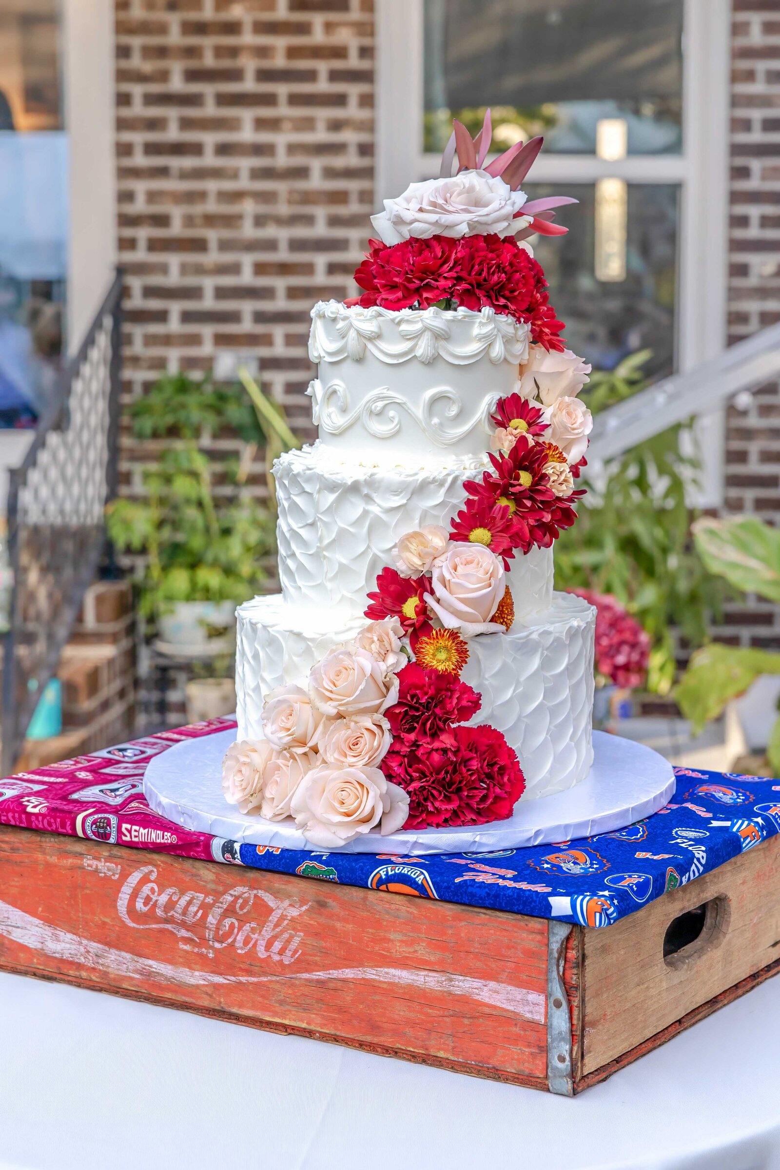 A three-tiered wedding cake decorated with red and blush flowers, displayed on a vintage Coca-Cola crate, captured during a Wilmington, NC wedding, showcasing rustic charm and creative details.