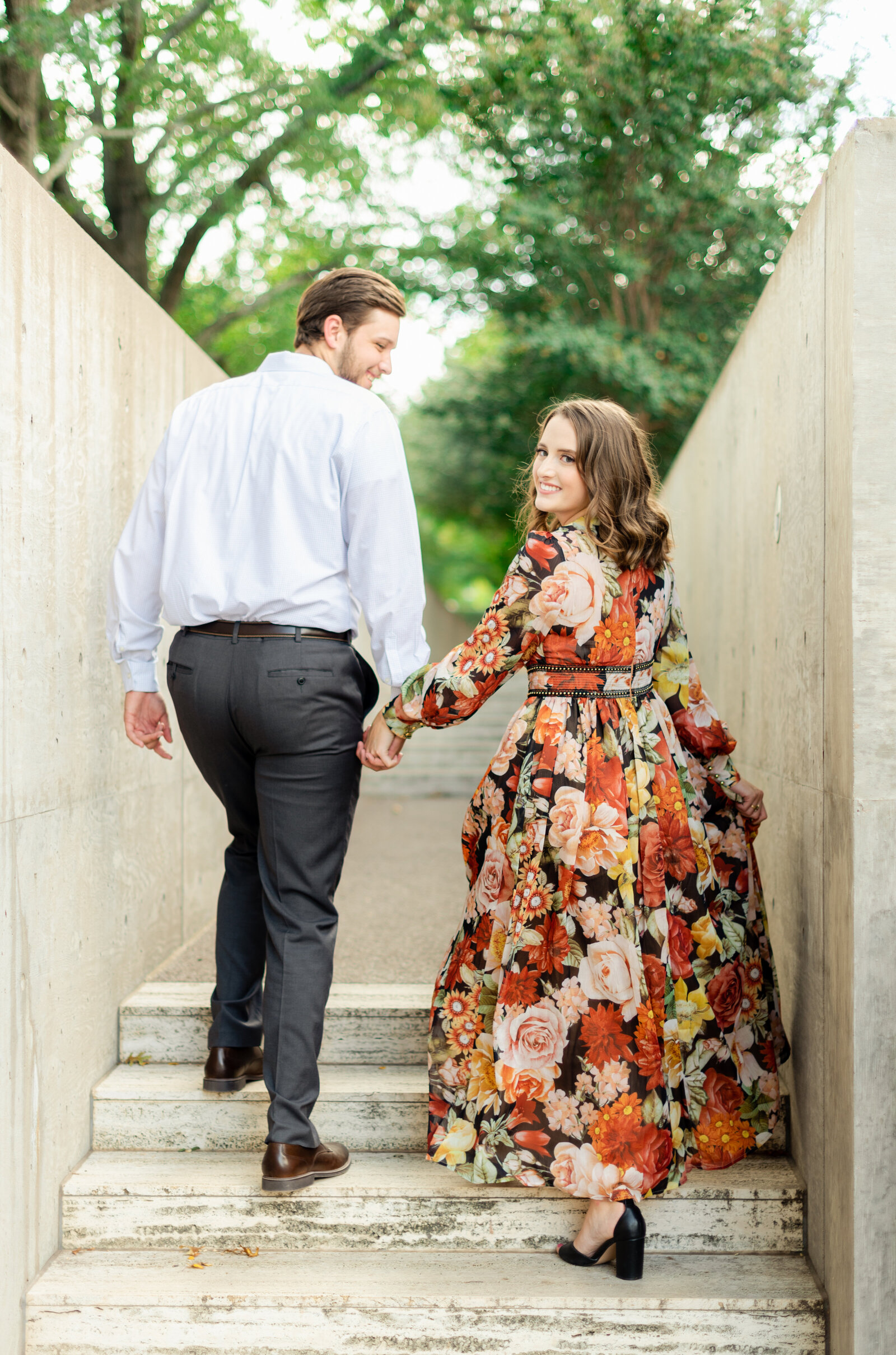 Portrait of a man in a white shirt and gray dress pants holding hands with a woman in a floral dress on the stairs.