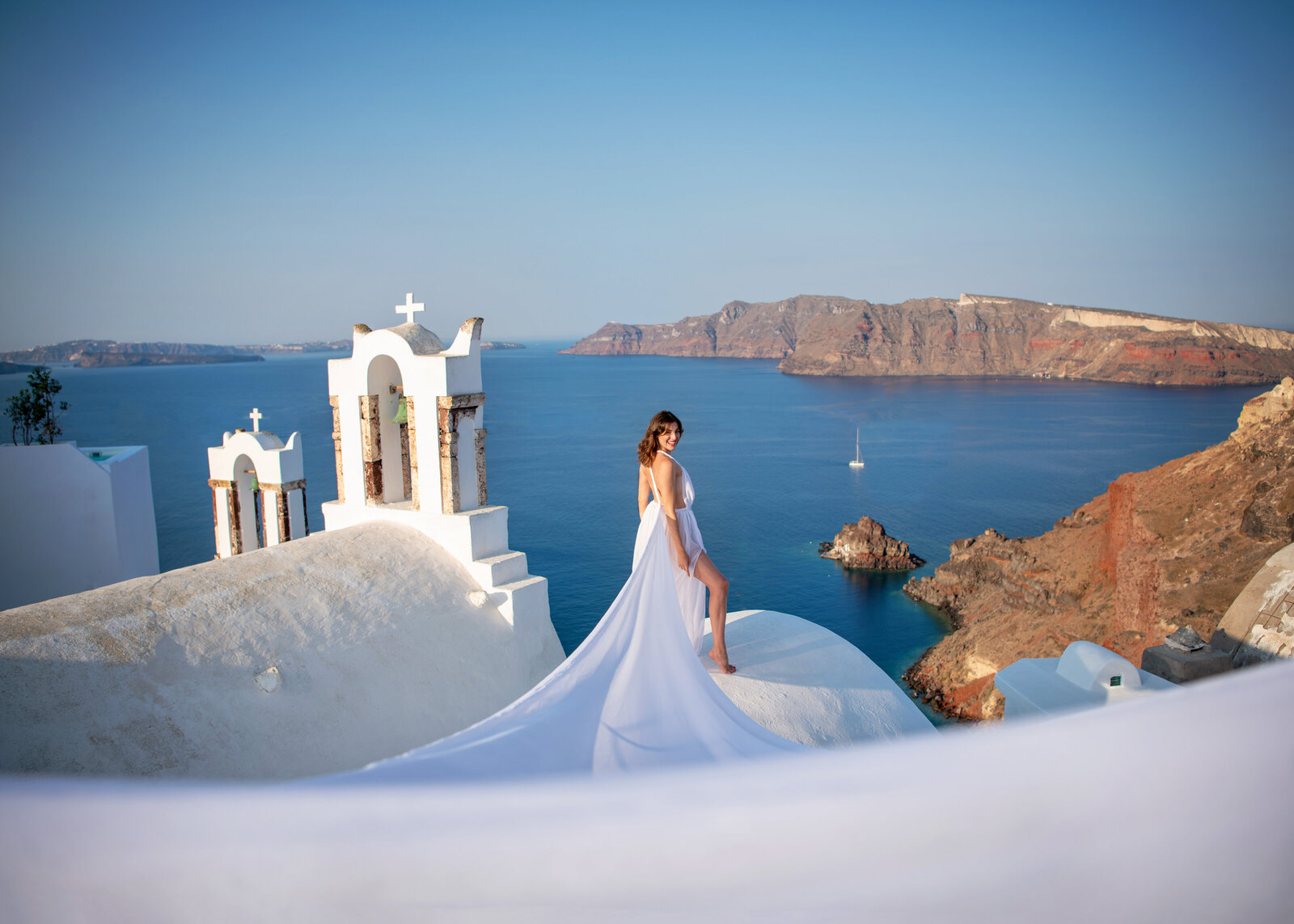 Adventurous destination bride in a wedding gown with a long train standing on the white rooftops next to a church in Santorini, Greece for destination shoot