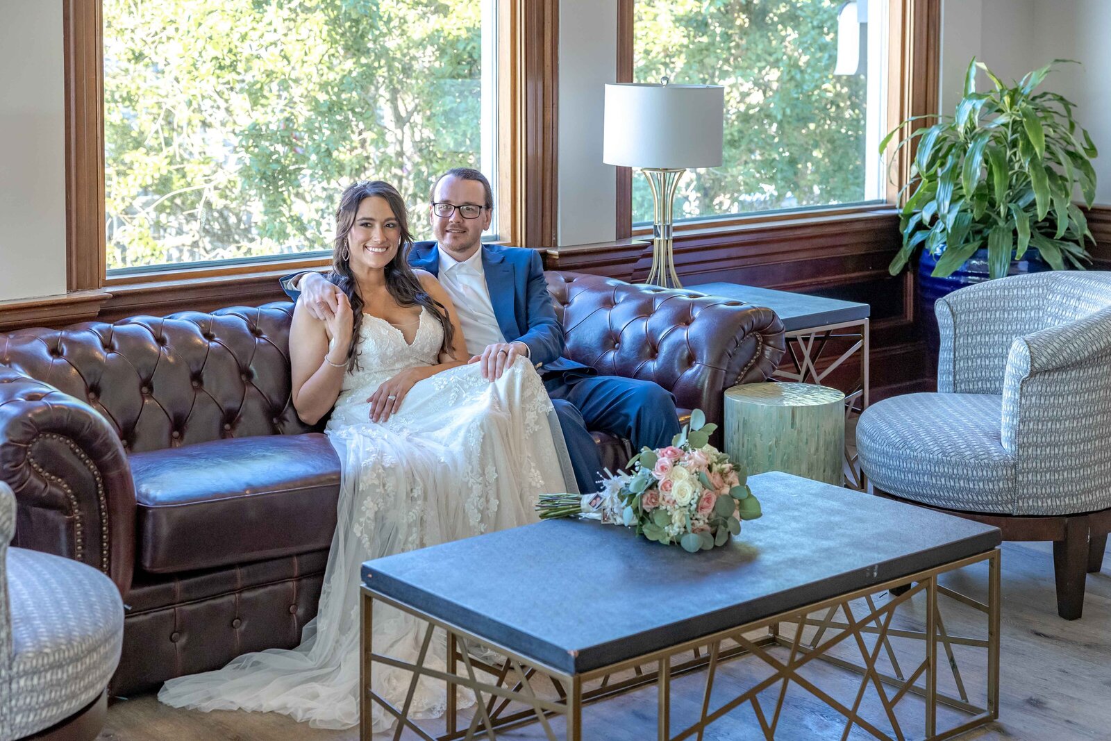 The bride and groom relax on a vintage leather couch in a bright and elegant venue, surrounded by natural light and greenery, captured during a Boone, NC wedding, showcasing rustic elegance and romantic moments.