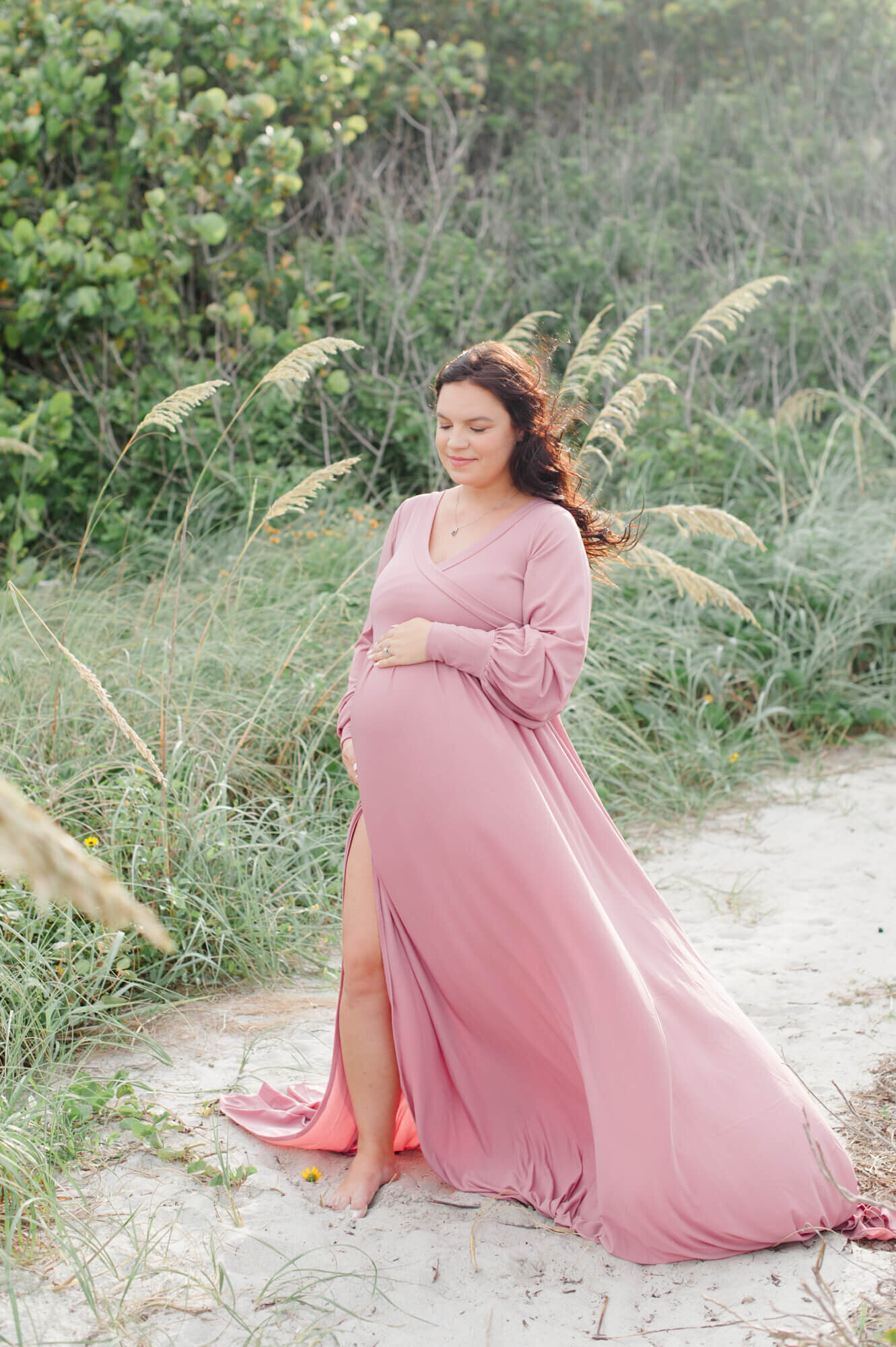 Mom standing near the dunes in a gorgeous pink gown holding her belly during her maternity photoshoot