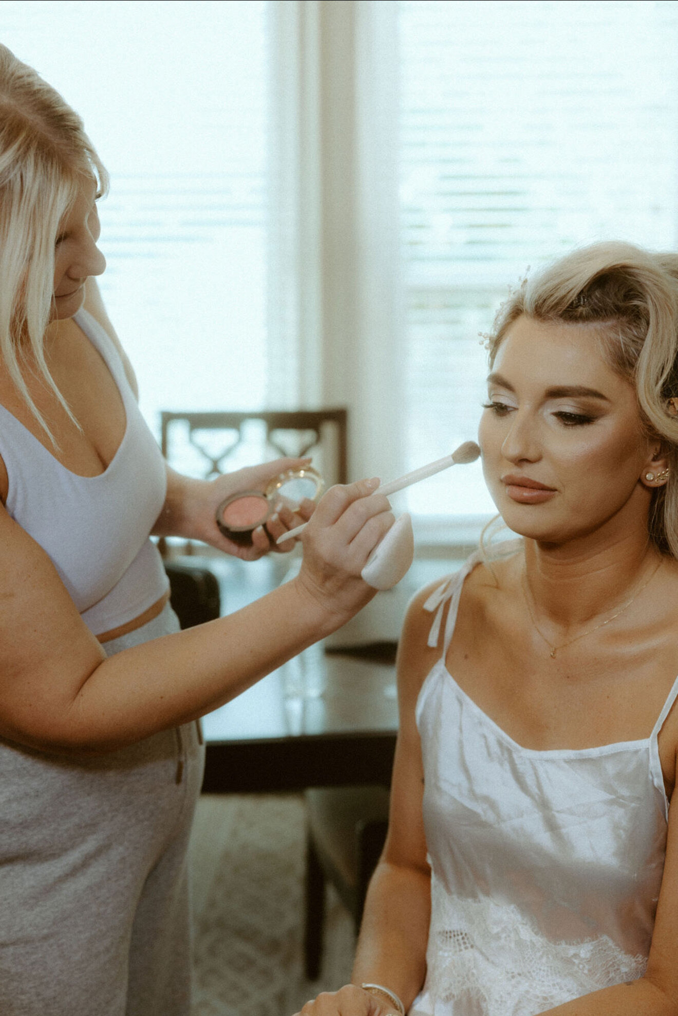 A woman in a white dress is getting her makeup done by an editorial bridal makeup artist, showcasing modern bridal makeup trends in a brightly lit room.