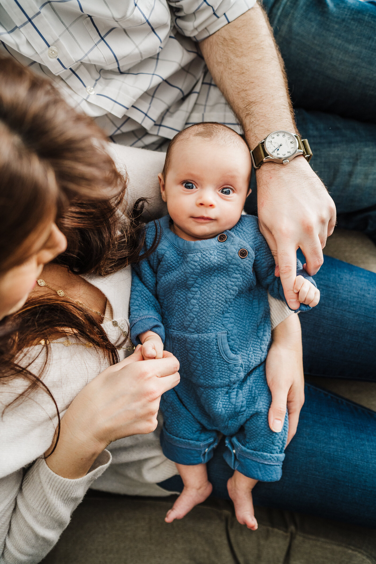 baby boy with blue eyes looks at camera wearing a blue romper and laying in his parents laps