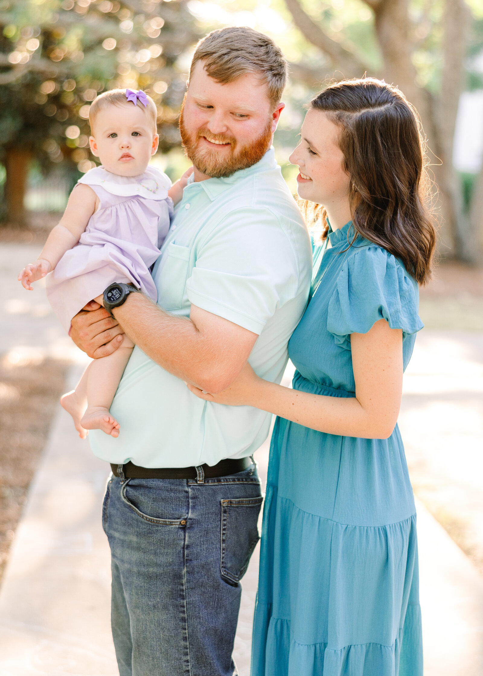 Father holding baby in a lilac dress while mom smiles lovingly, photographed by Abby Bell Photography, Fine Art Portrait Photographer based in Alabama.