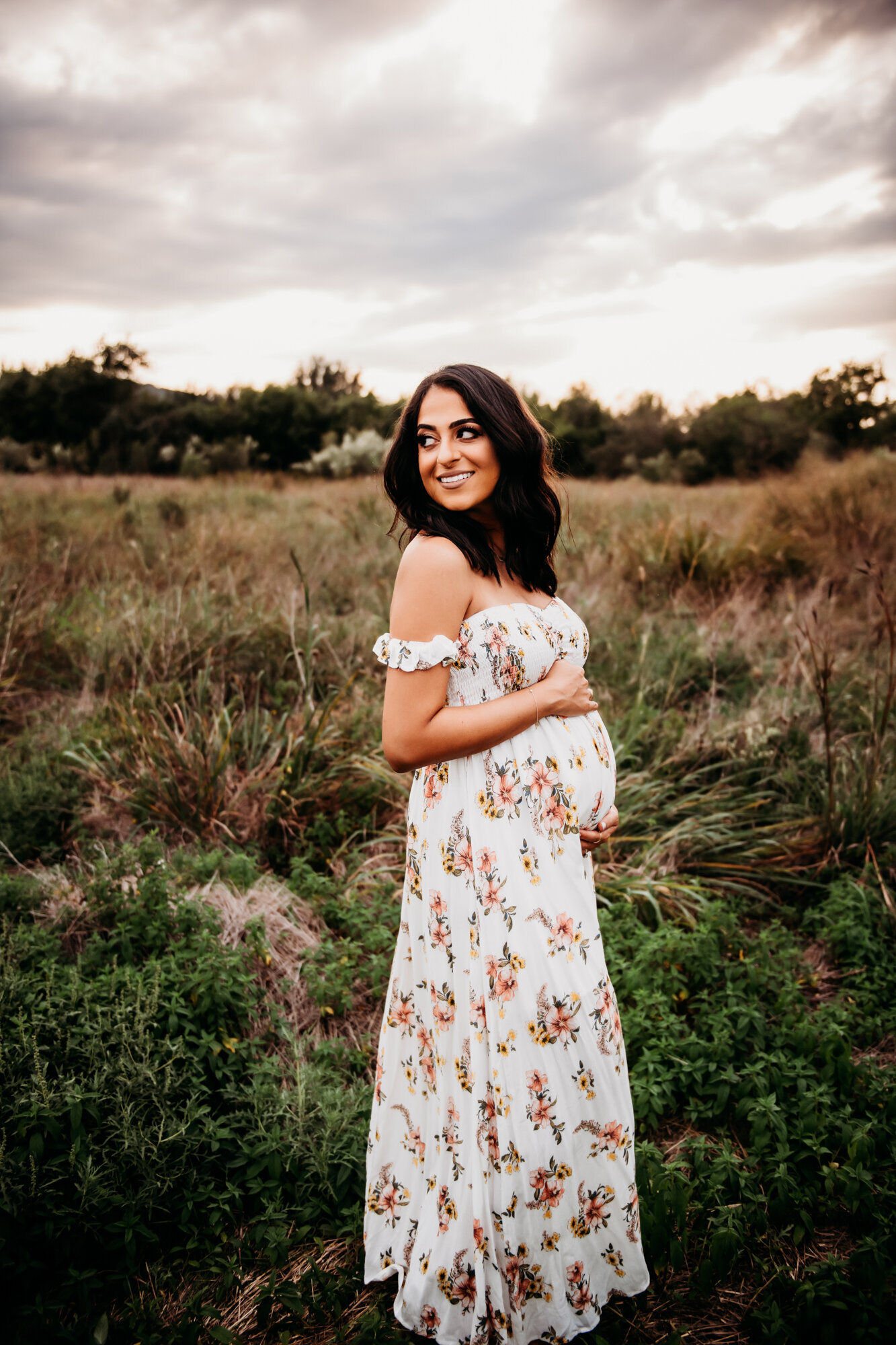 Maternity Photographer, A pregnant woman in a floral dress is standing in a field as she looks over her shoulder and smiles.