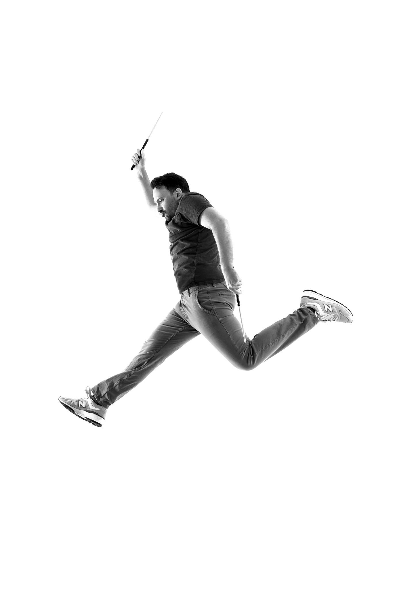 A man in casual clothing jumps high in the air with legs spread wide and one arm raised, holding a conductor’s baton, against a white background—a dynamic moment captured by a skilled Jacksonville band photographer.