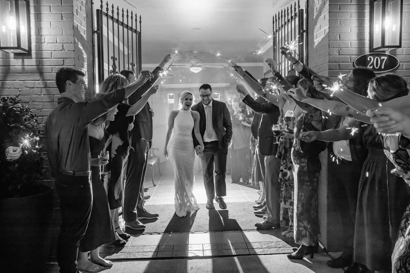 A bride and groom walk hand in hand under an arch of sparklers held by cheering guests, celebrating their wedding at night. The scene is joyful and captured in black and white.