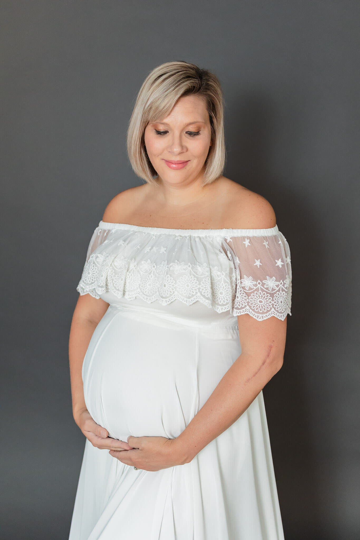 A pregnant woman wearing a white dress posing on a gray backdrop.