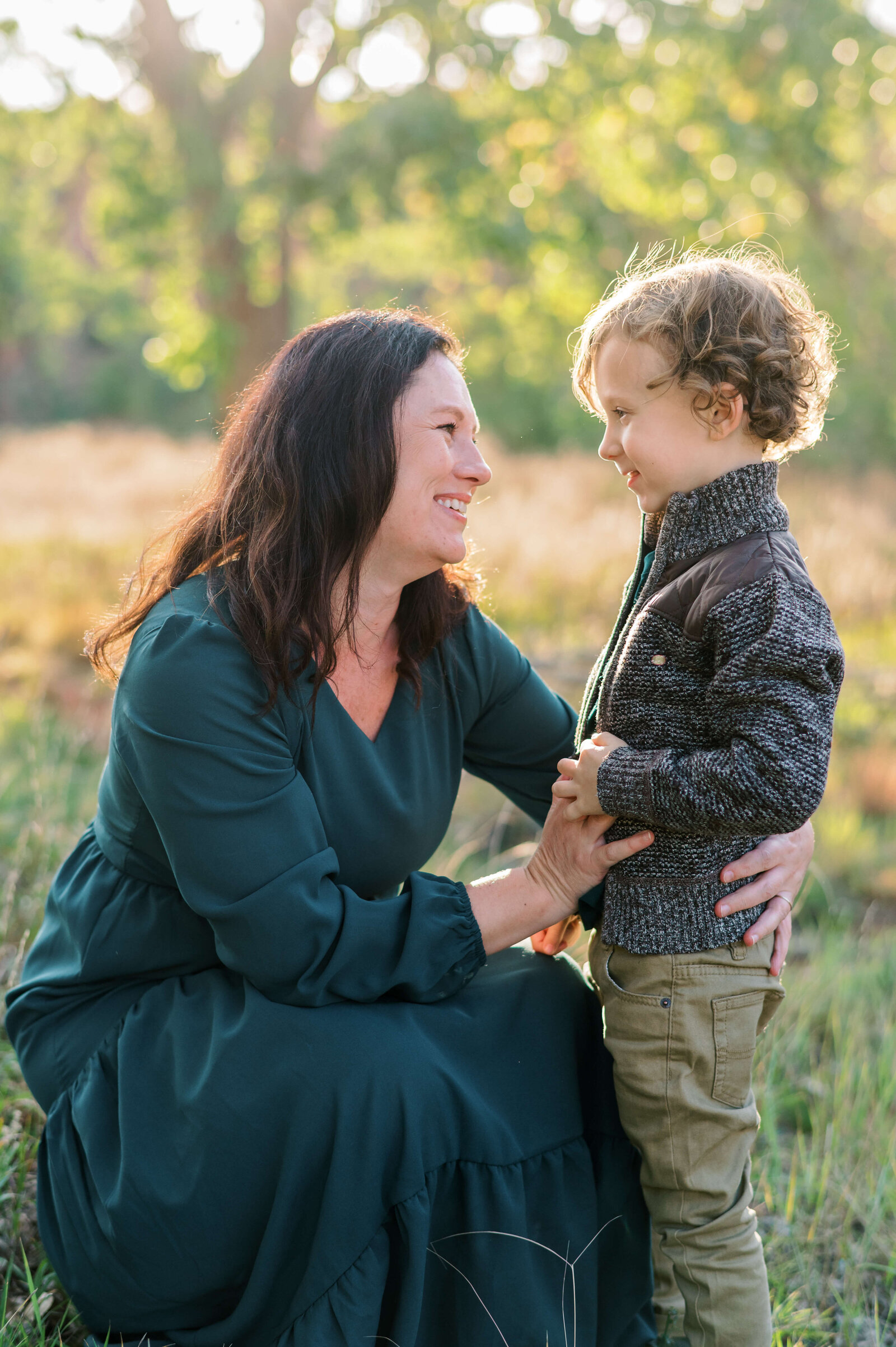 A brunette mother in a long green dress smiles at her young son who wears a tweed jacket and tan pants