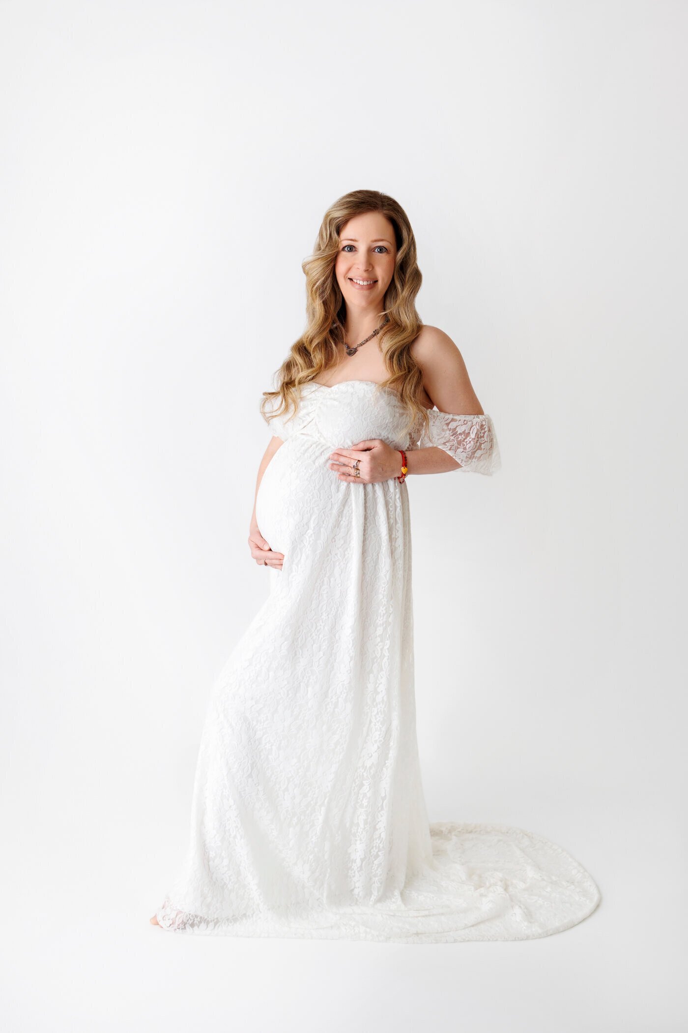 A pregnant woman stands gracefully against a white background, wearing an off-the-shoulder, lace-covered white gown that flows to the floor with a slight train. Her long, wavy hair cascades over her shoulders, and she smiles warmly at the camera while cradling her baby bump with both hands. The clean, minimalist setting emphasizes the elegance and joy of the maternity portrait.