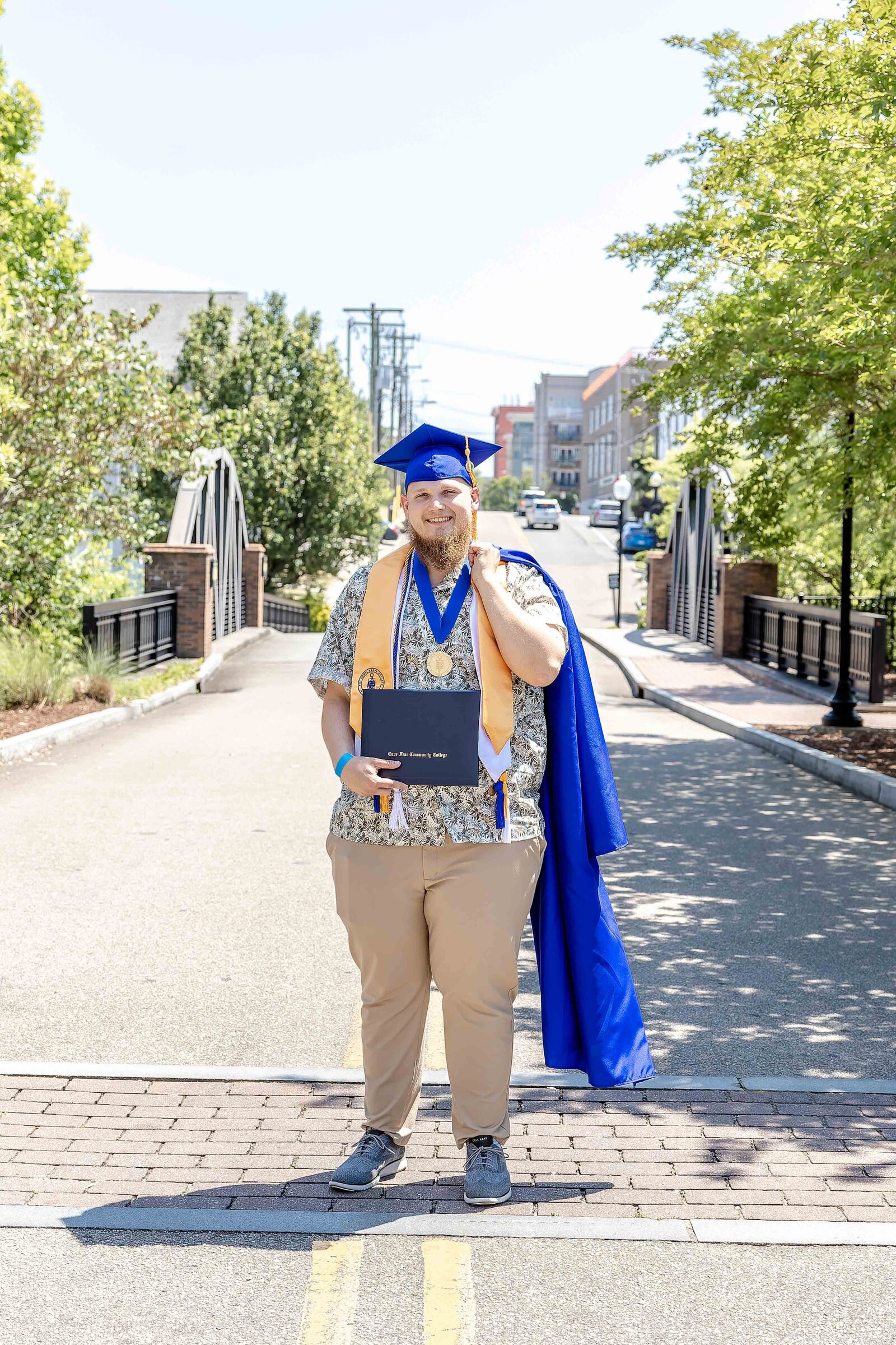 A Cape Fear Community College senior in a blue cap, gown thrown over his shoulder, and holding a diploma is standing confidently on a bridge in downtown Wilmington, North Carolina. This photo captures graduation excitement, ideal for vibrant and professional senior photography in an urban setting.