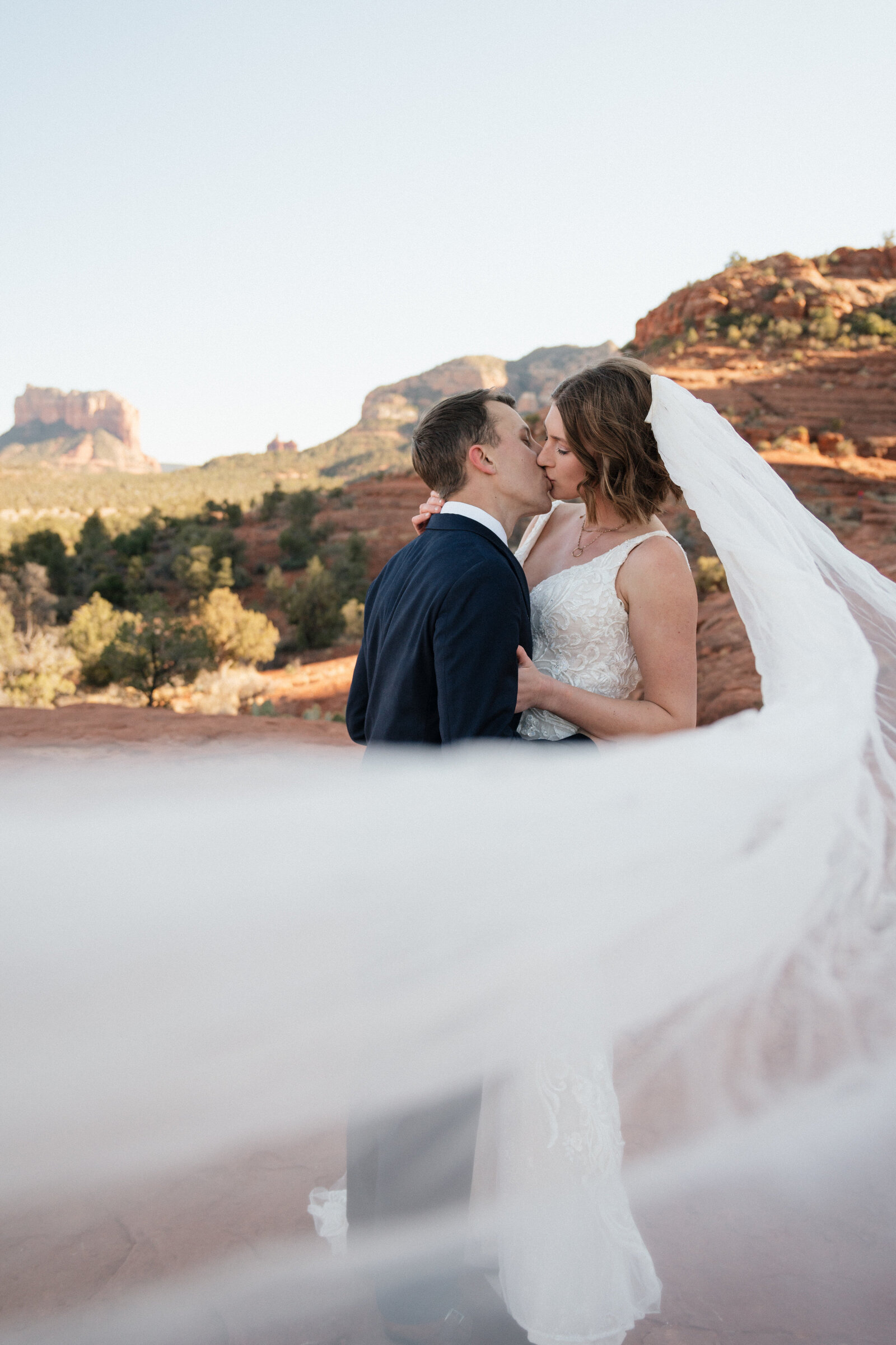 Bride and groom sharing a kiss with the bride’s veil flowing in the wind during their Cathedral Rock elopement in Sedona, surrounded by red rock views.