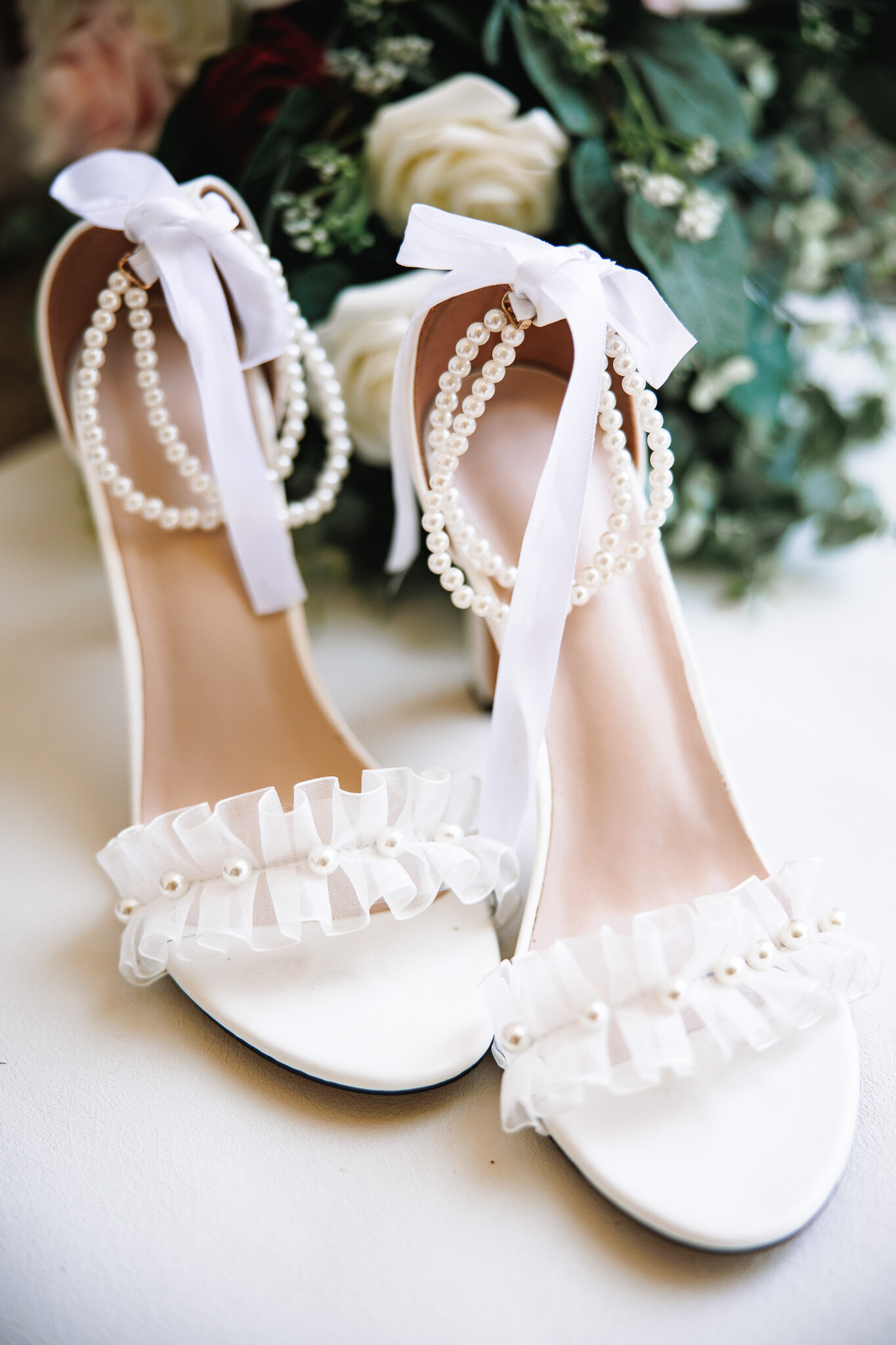 A detail photo of a pair of white strappy heels detailed with lace, ribbon, and pearls.