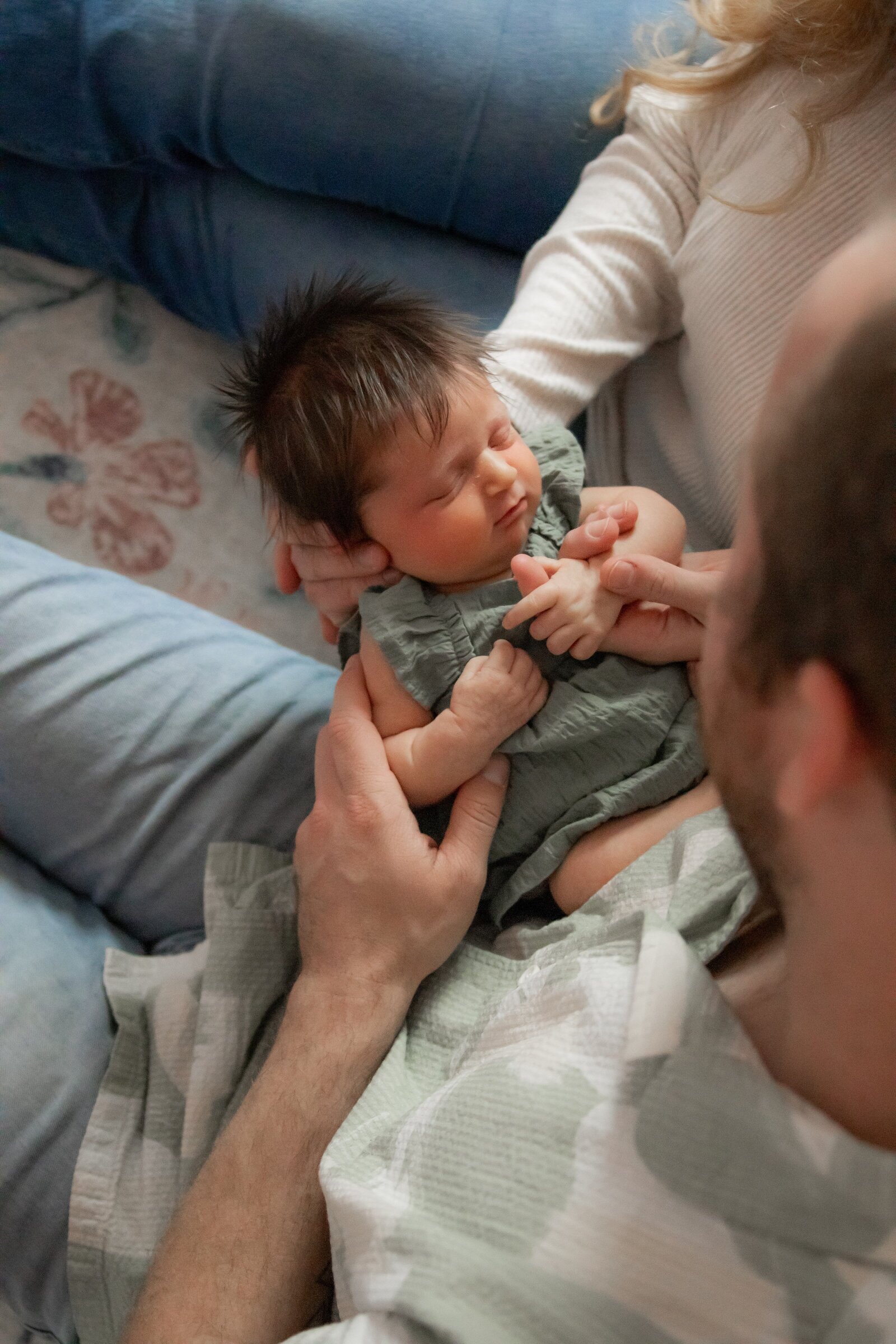 Newborn baby girl in focus looking down while mom and dad sit on floor in front of crib looking down at their baby.