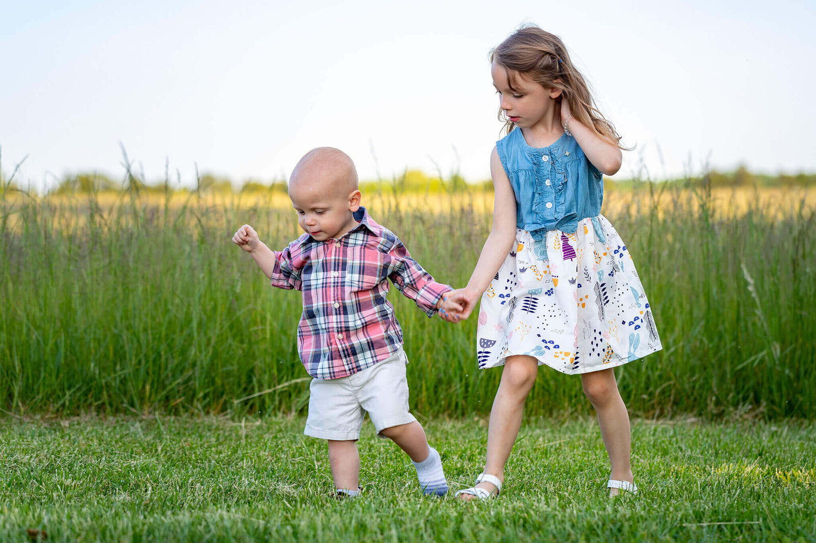 A brother and sister holding hands and walking in the grass.