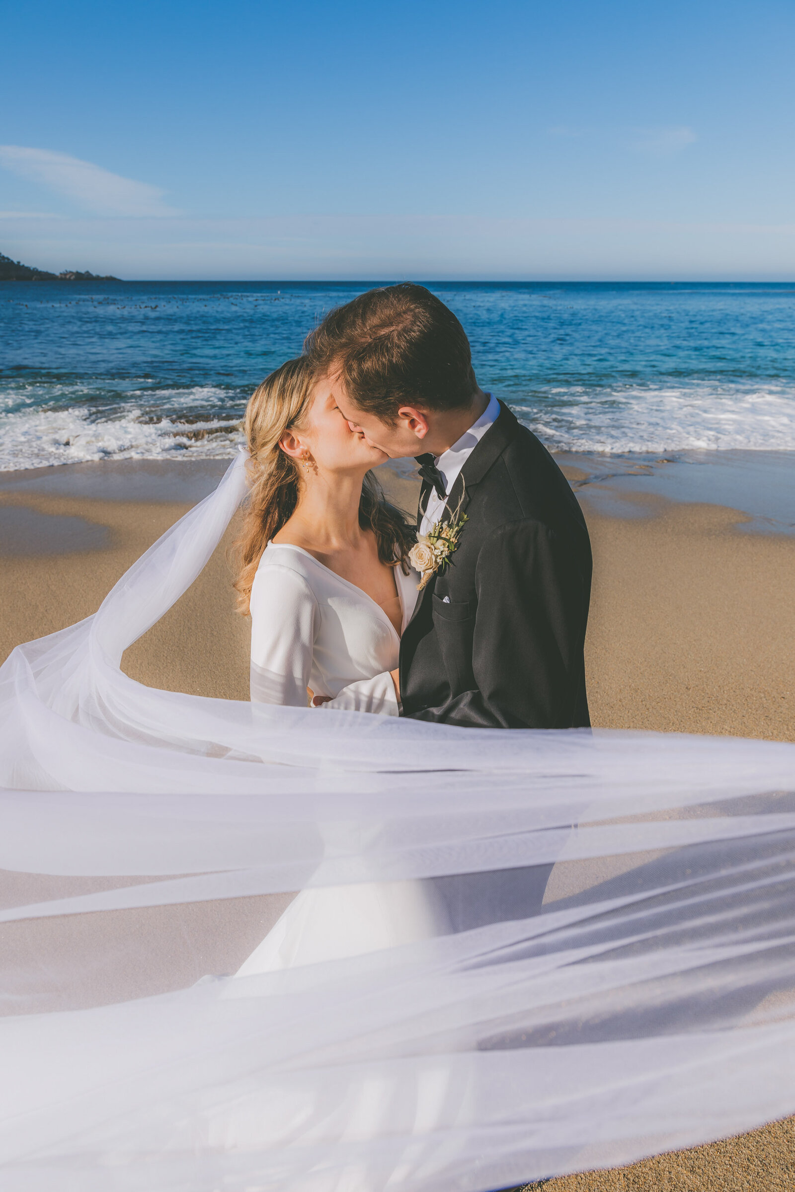 A bride and groom kiss on a beach while her veil is carried in the wind and wraps around the bottom of the photograph.