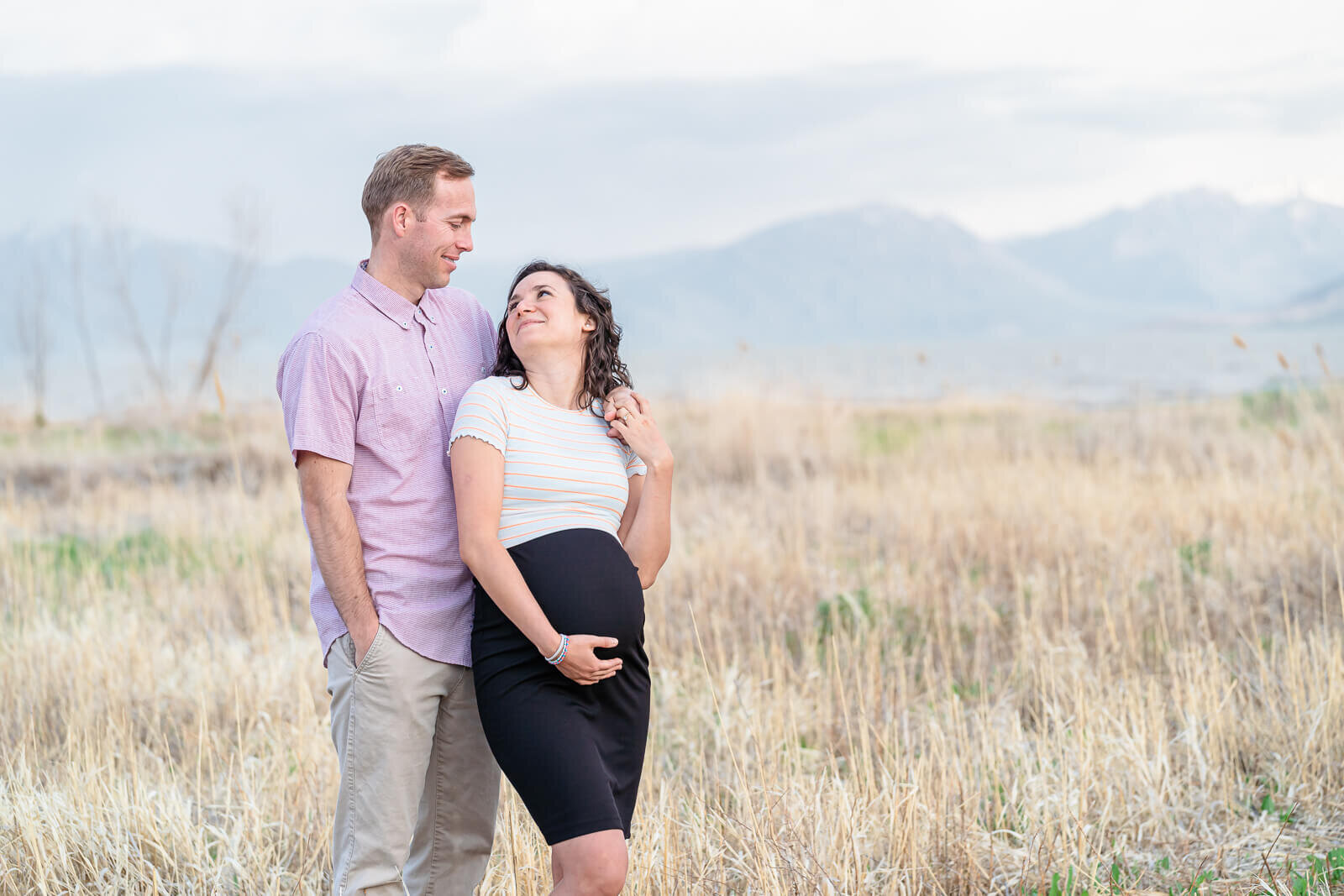 a pregnant woman wearing a white striped shirt and black skirt with her husband standing behind her. Captured by Utah Lake in Lehi