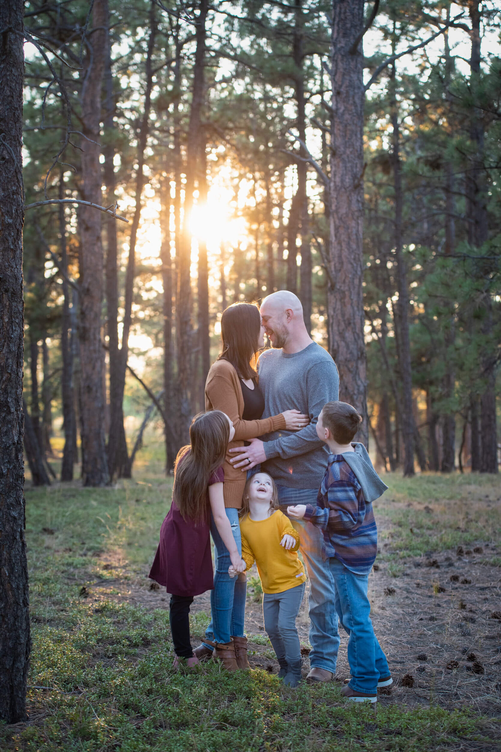 Mom and dad kiss while their three toddlers play around them looking on as posed by a Denver family photographer