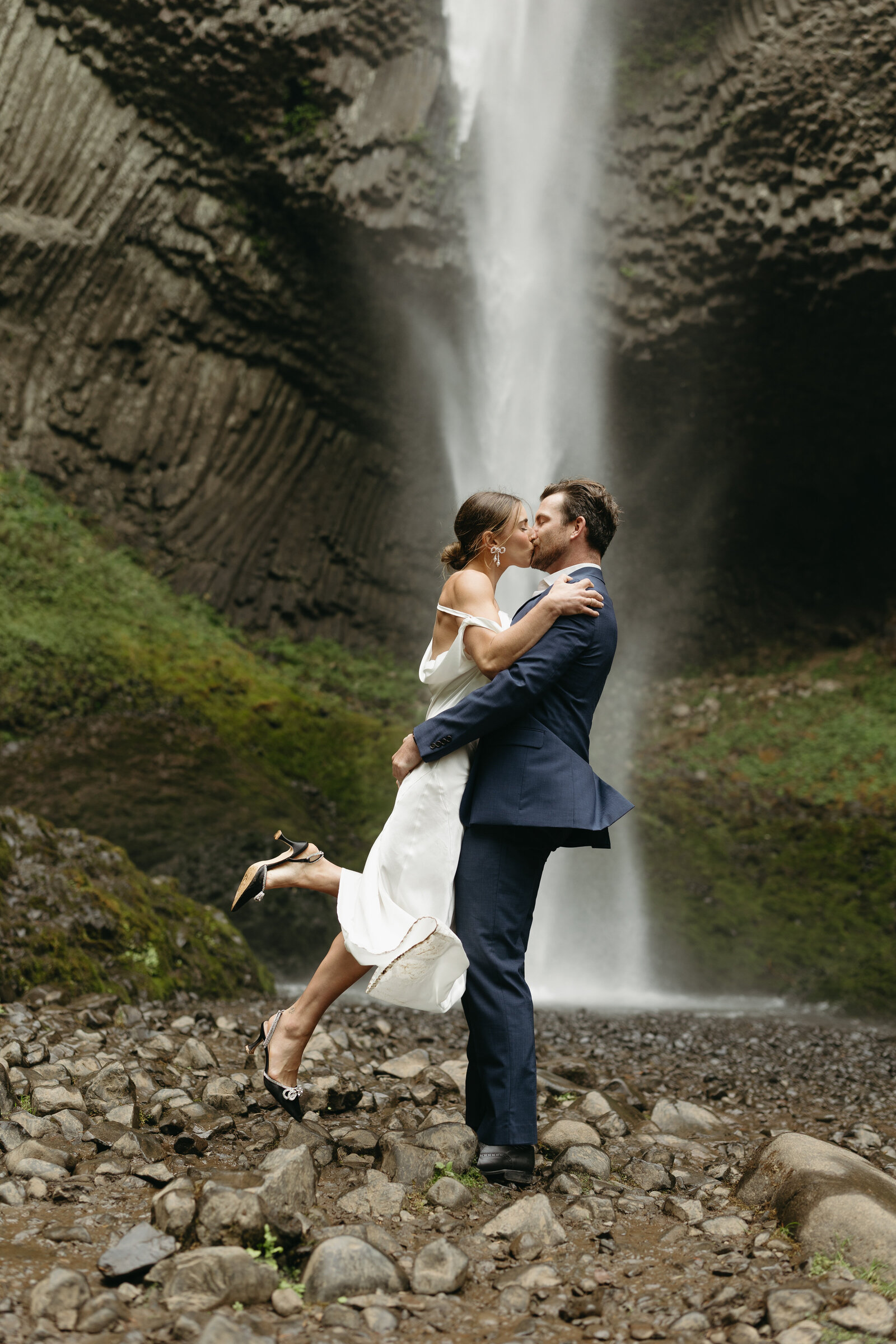 A groom is lifting and kissing his new wife at a waterfall after their Oregon elopement. 