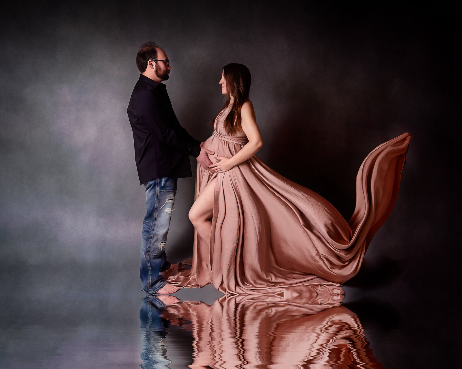 A father-to-be tenderly holds hands with his pregnant partner, who wears a billowing blush gown, symbolizing love and anticipation.