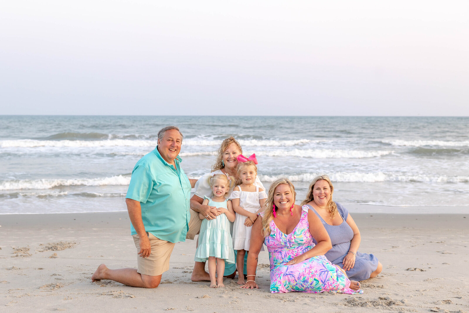 A multi-generational family poses together on the beach, dressed in colorful summer attire. The children and adults share smiles against the ocean backdrop, ideal for professional family photography in NC, highlighting warm family bonds.