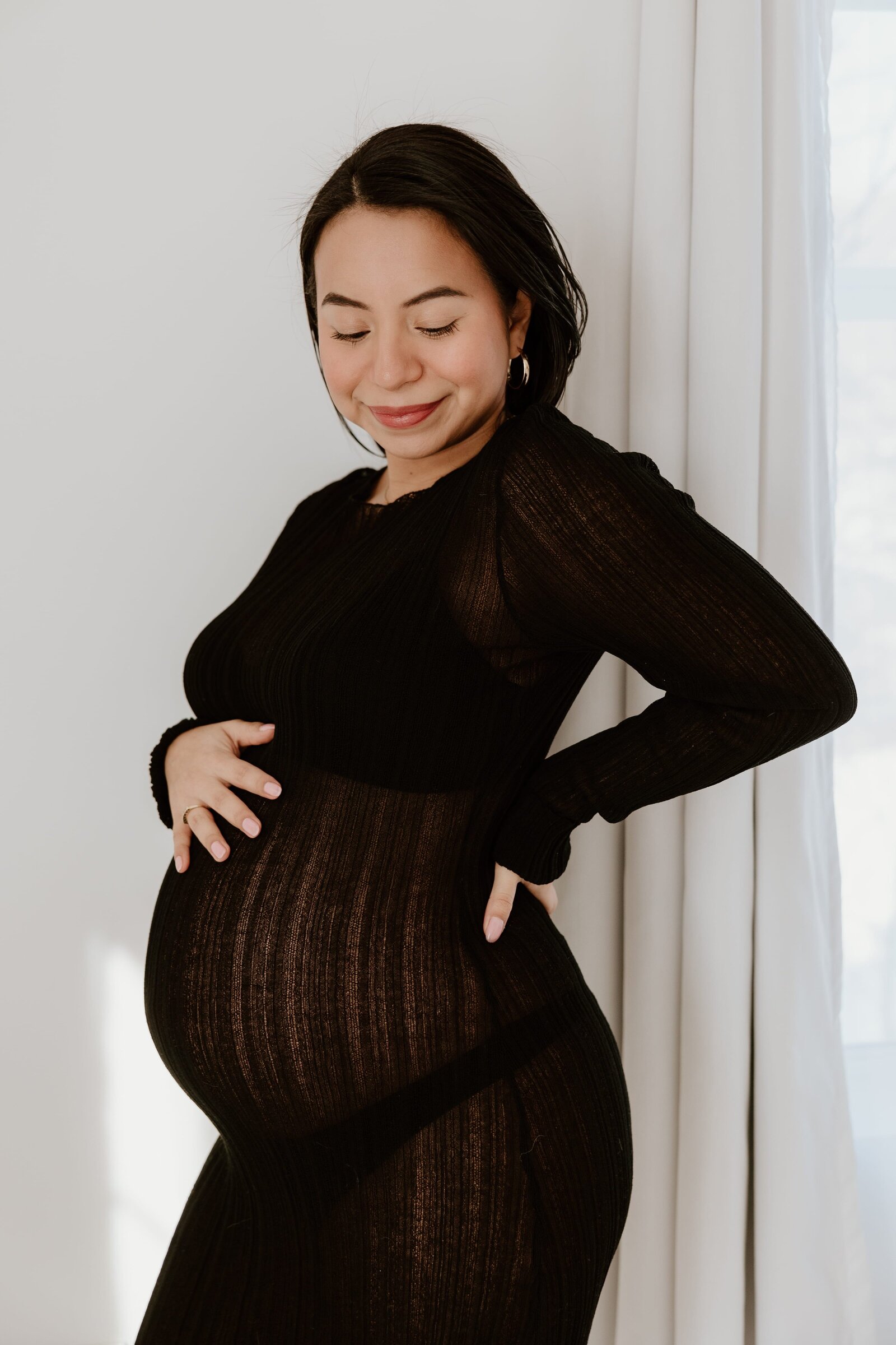 Beautiful maternity portrait of a smiling woman in a sheer black dress, cradling her baby bump with soft natural light in the background.
