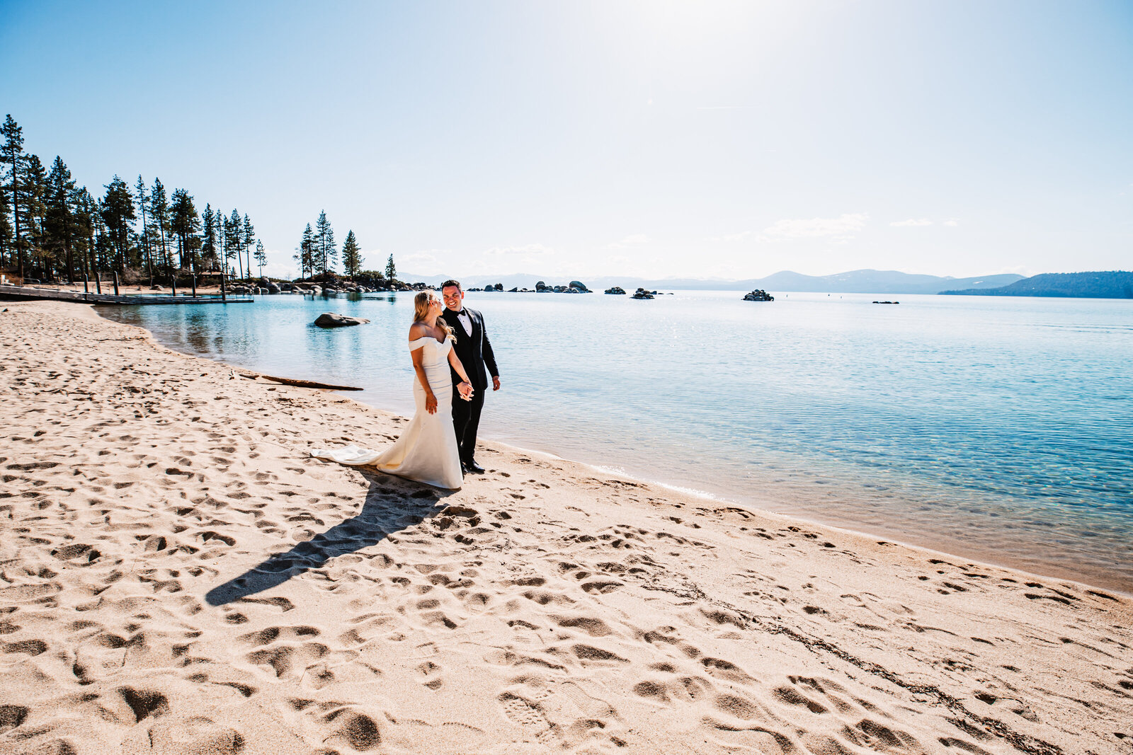 A newlywed couple walks hand in hand along the sandy shore of Lake Tahoe, soaking in the crystal-clear waters and mountain views on their elopement day.