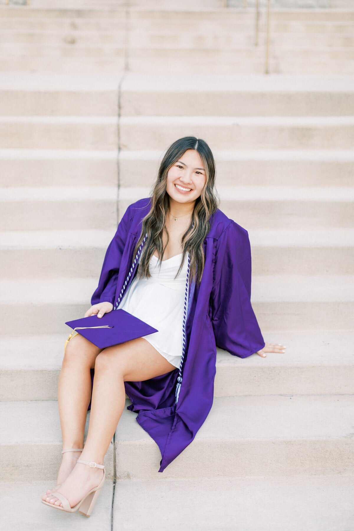 An asian girl sits on the steps at Wilson Hall, and smiles as she wears her purple and gold cap and gown.