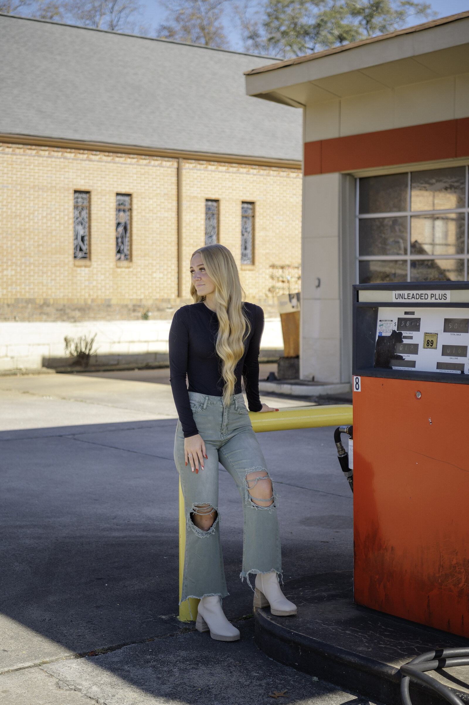 Senior with long blonde hair stands beside a red gas pump at an old gas station