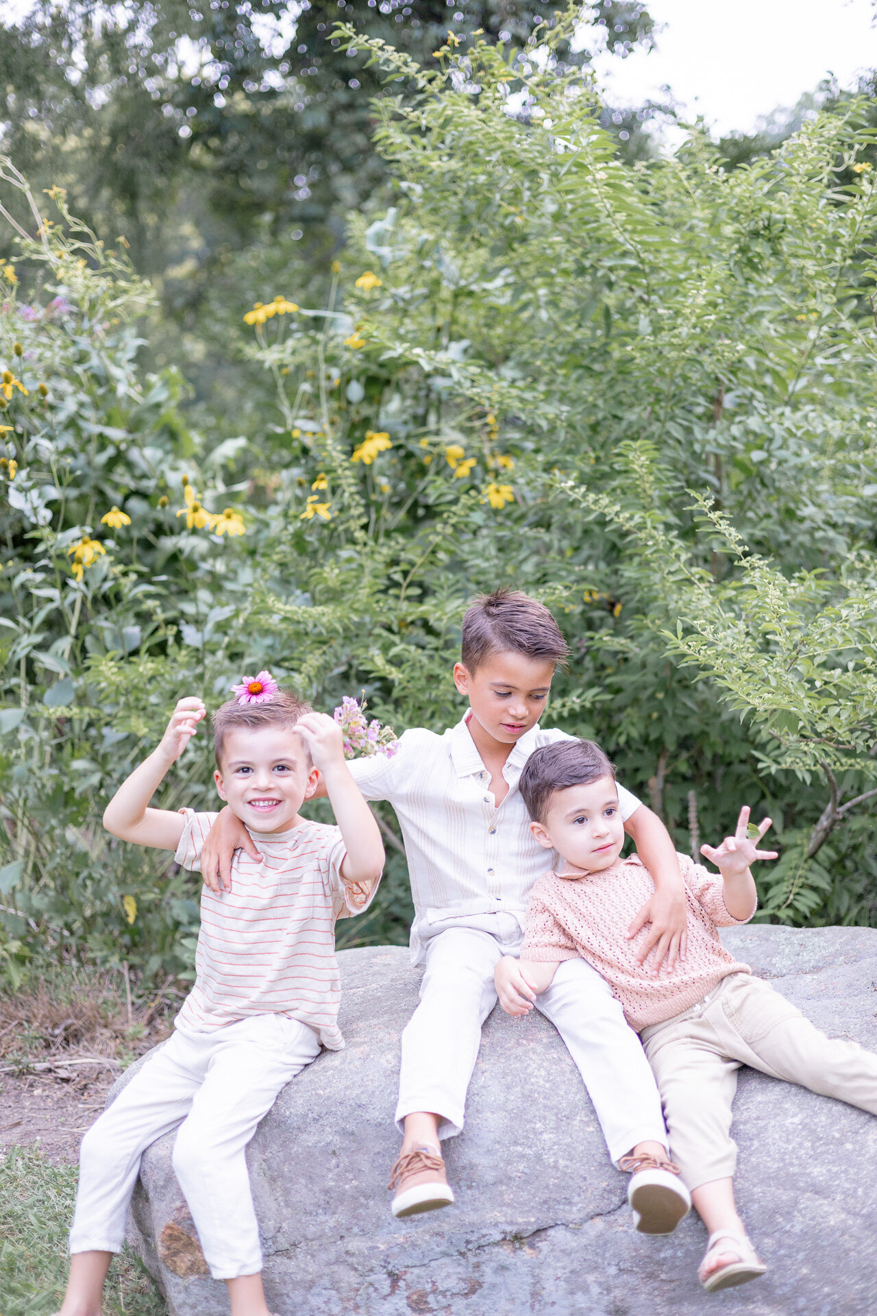Three young brothers sitting on a large rock surrounded by greenery and wildflowers, captured during a lighthearted family photo session in Connecticut. One boy wears a flower crown, while the others share a silly moment outdoors.