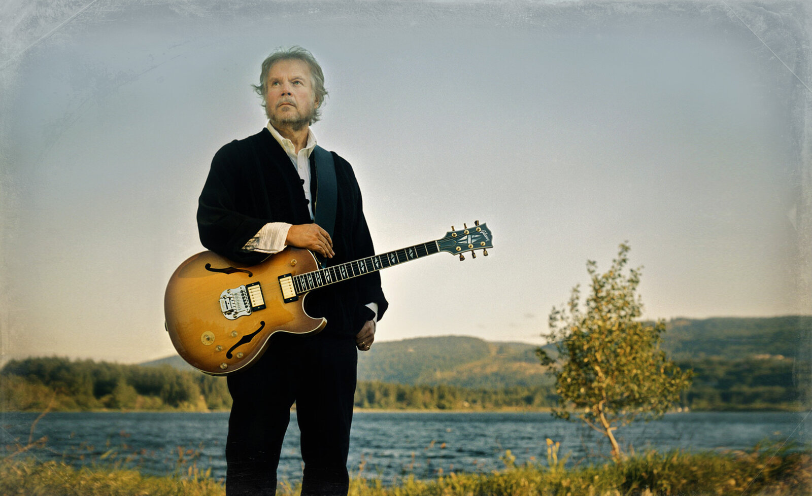 Randy Bachman portrait standing with guitar wearing black suit lake and small tree behind