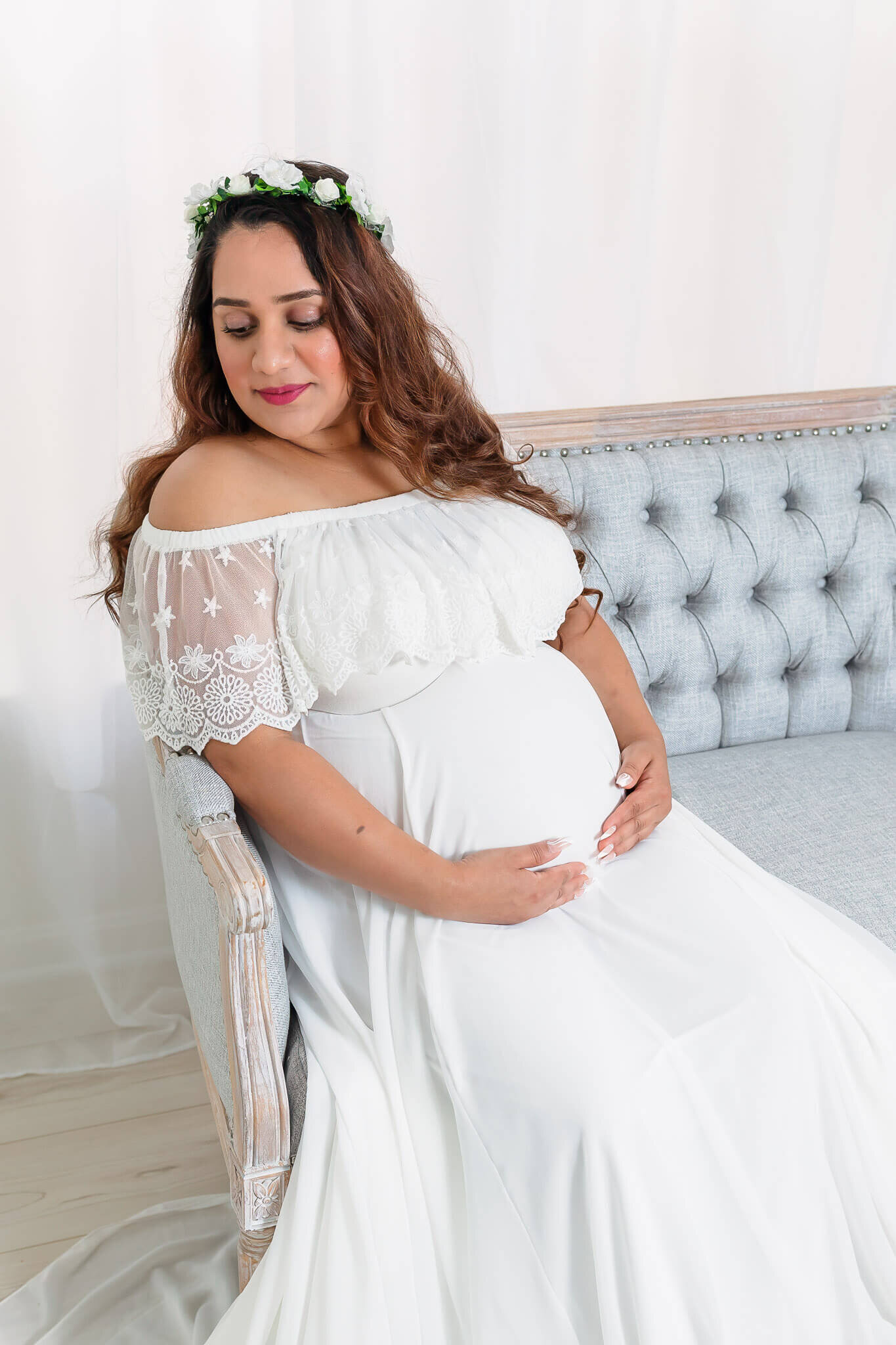 A mom-to-be in a white dress sitting on a couch in a Gainesville studio.