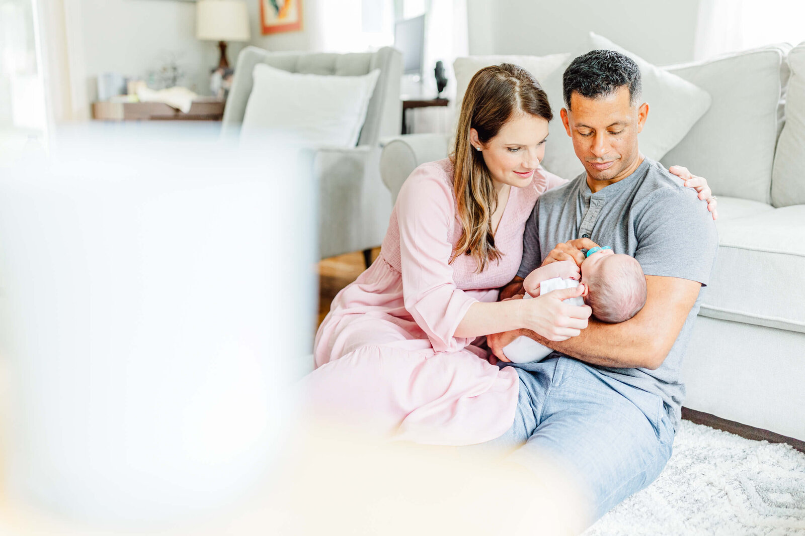 Pulled back photo of mom and dad sitting on the floor interacting with their newborn