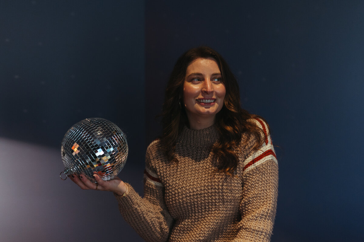 A woman in a sweater holds a disco ball, subtly smiling, symbolizing corporate event planning creativity in Northwest Arkansas.
