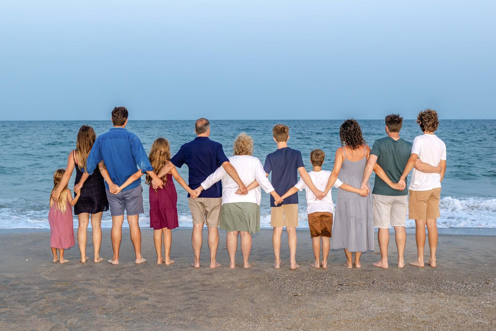 A family stands hand in hand, facing the ocean on a sandy beach. Dressed in a mix of casual summer outfits, they create a united silhouette against the serene water. Perfect for North Carolina family photography, capturing togetherness and tranquility.