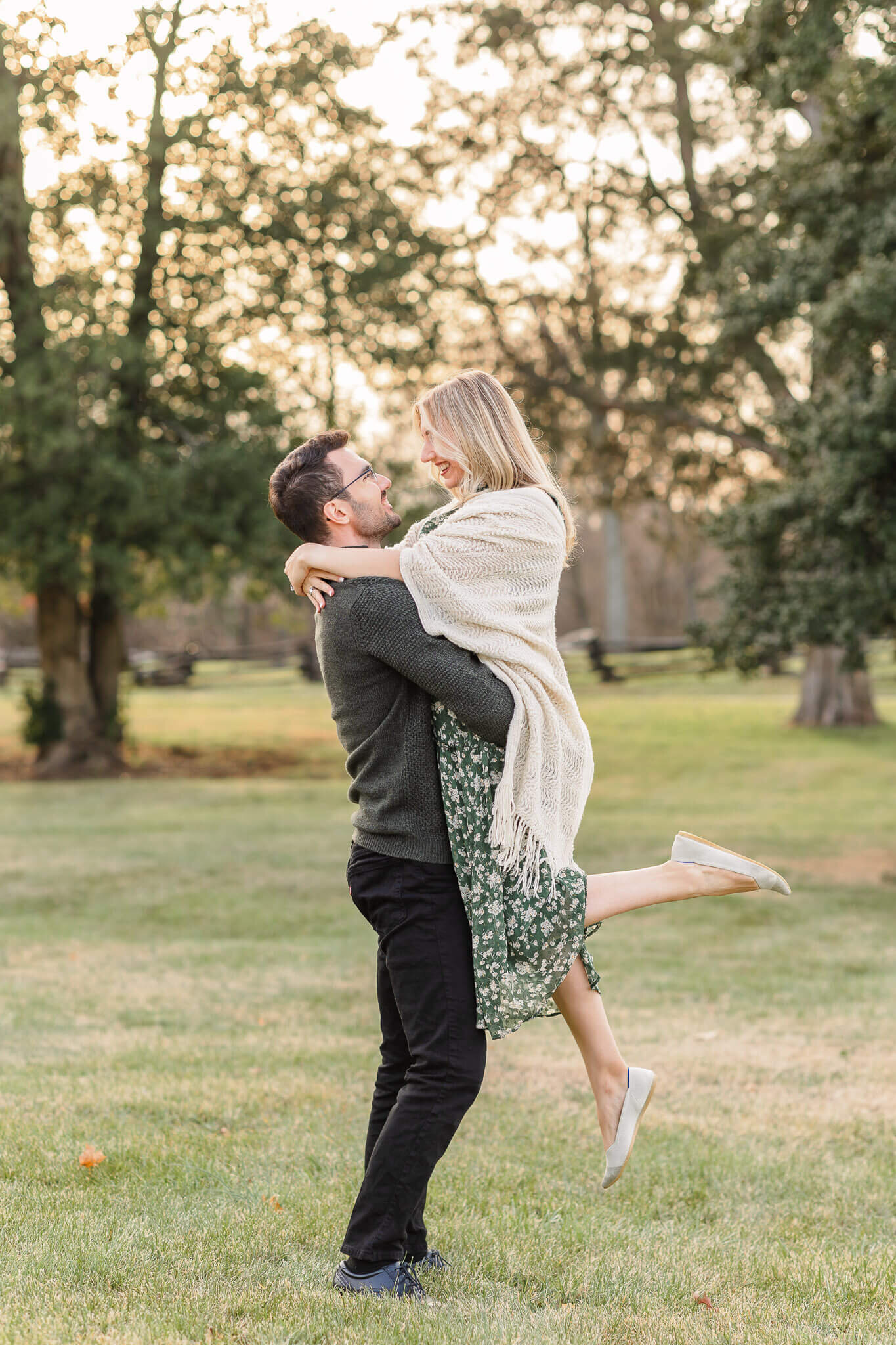 A dad-to-be lifting his wife into the air at a park at sunset.