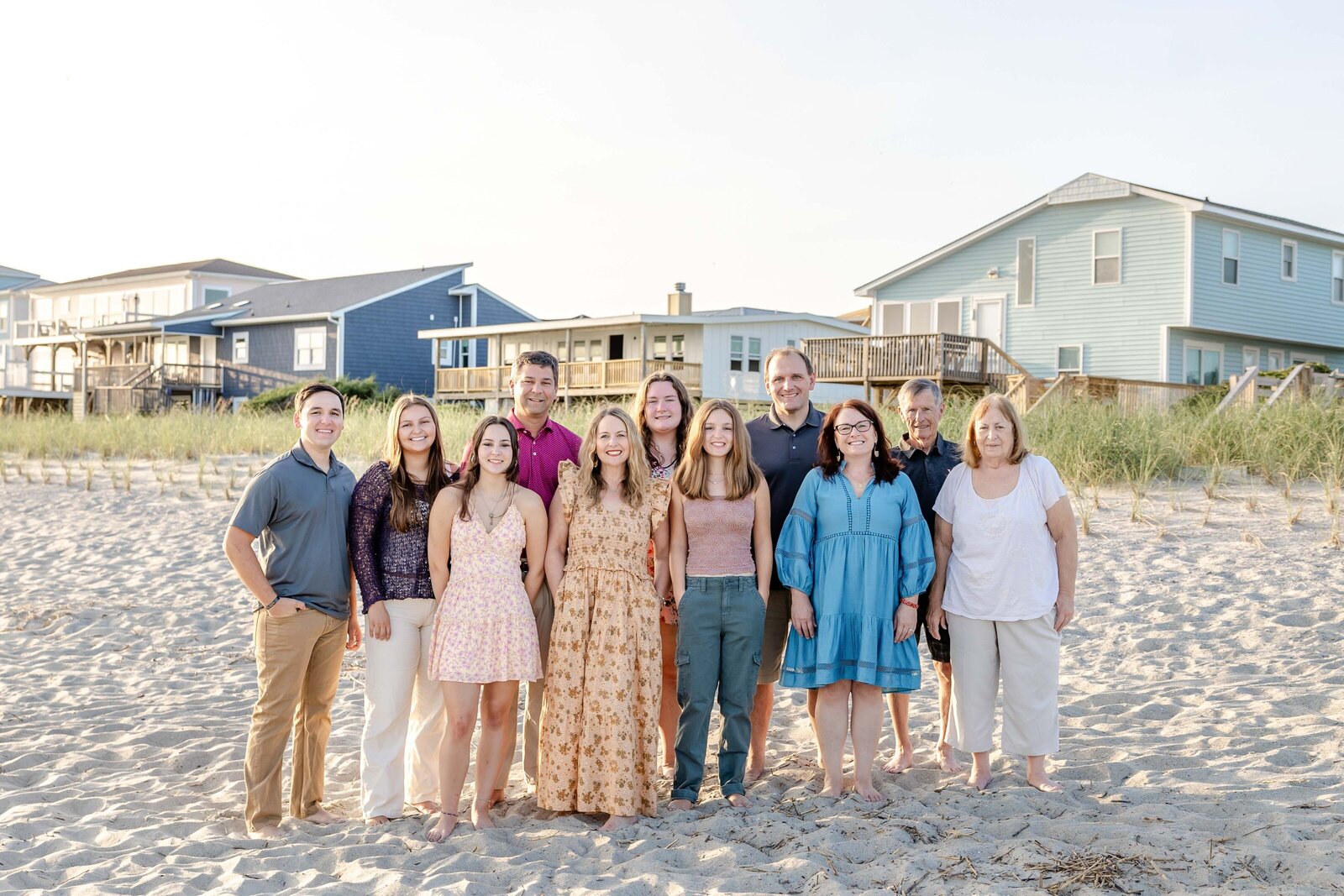 A family stands on the sandy beach in front of colorful coastal homes. Dressed in casual summer outfits, they enjoy a sunny day by the sea, ideal for professional family photography in NC, showcasing vibrant beachside gatherings.