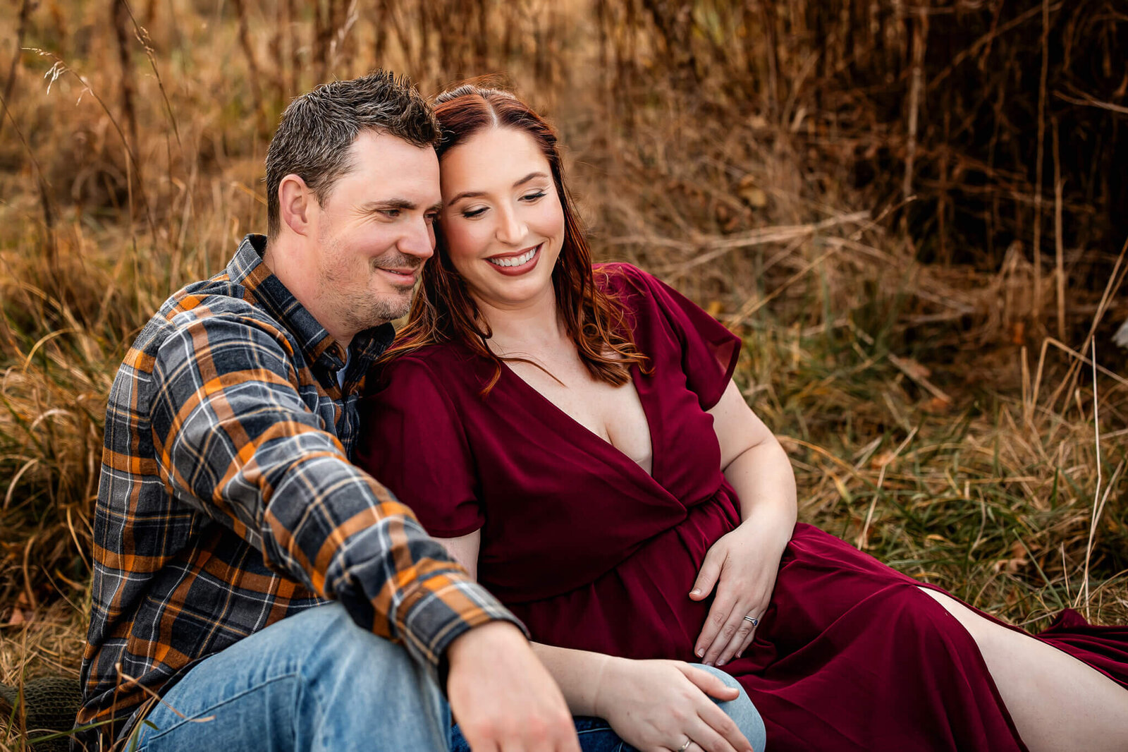 A smiling couple sits close together outdoors on grass for maternity portraits. The man wears a plaid shirt and jeans, while the woman, with long red hair in a burgundy dress, gently rests her hand on her belly amid brown, dried grass.