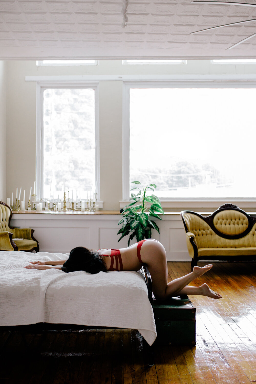 Brown hair girl dressed in a red lingerie set laying on a white bed with large windows captured by boudoir by Steph