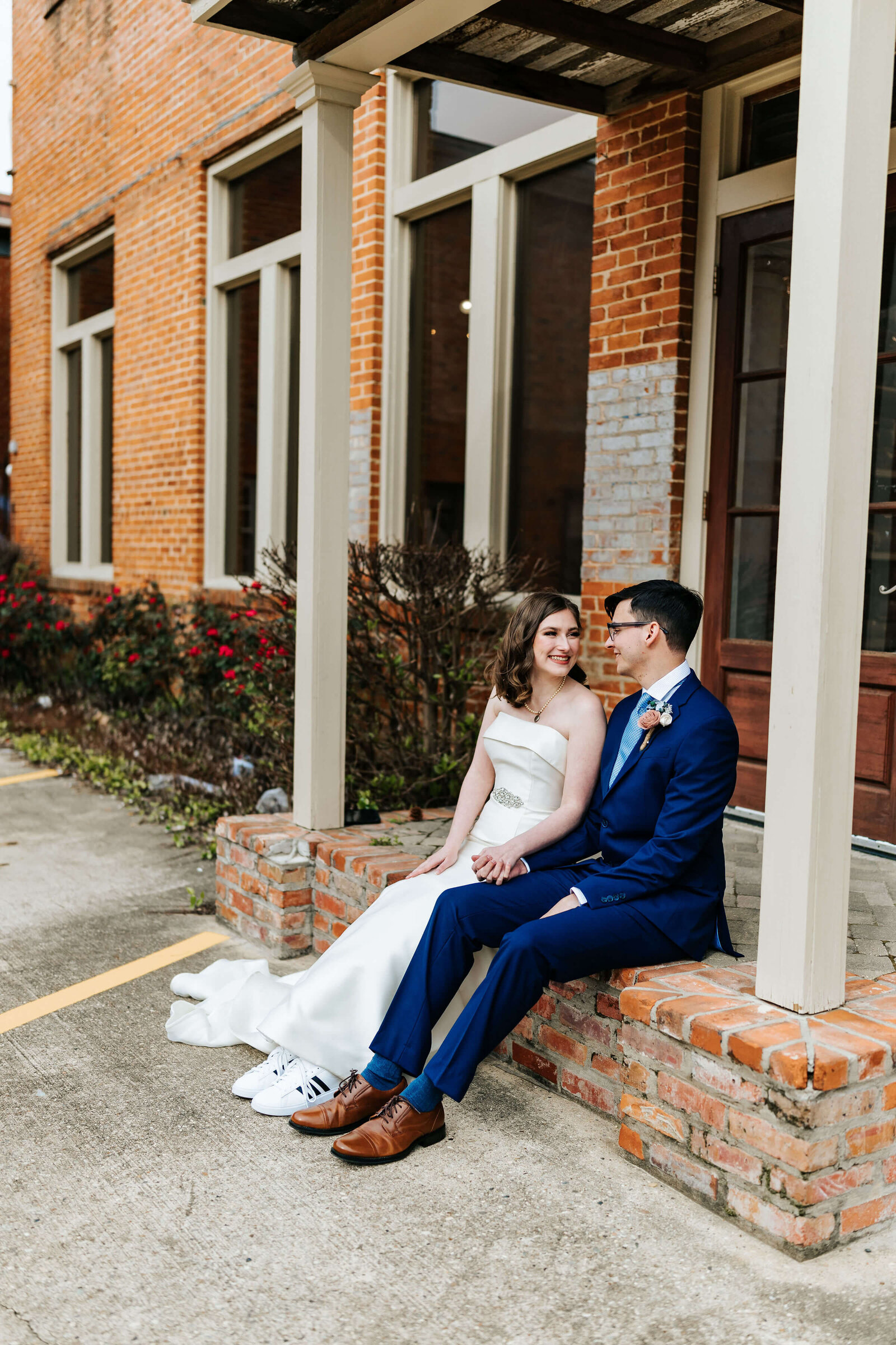 A bride in a white dress and sneakers sits beside a groom in a blue suit on a brick ledge outside a brick building. They are holding hands, smiling, and looking at each other.