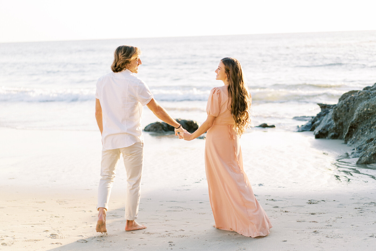 Portrait of a man in a white shirt and khakis holding the hand of a woman in a pink dress on the sand at the beach.