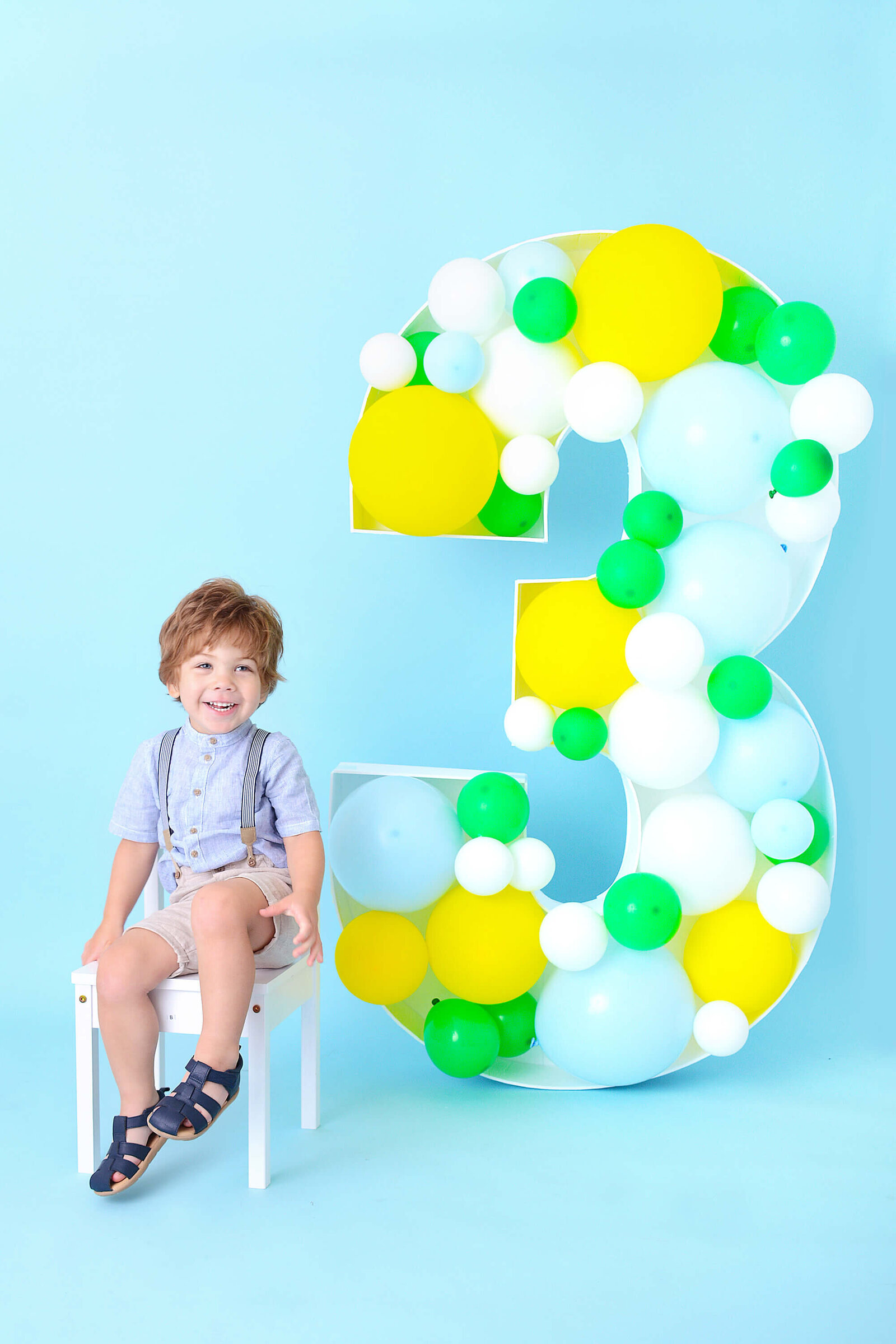 boy smiles with a large 3 made from balloons at his colorful birthday shoot