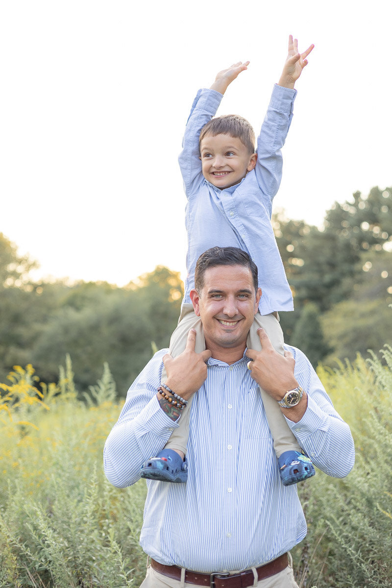 Smiling dad with his son on his shoulders during a golden hour family session in a wildflower field—captured by Westchester lifestyle photographer Nicole, known for relaxed and joyful family photography.